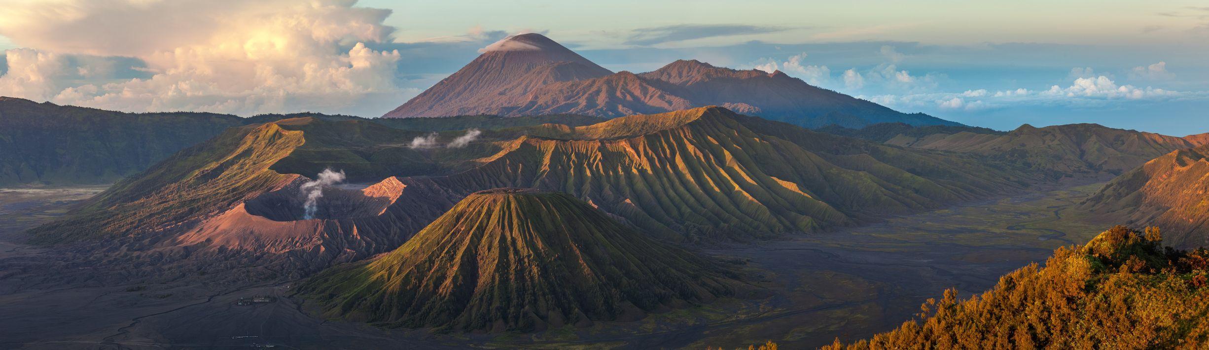 Ein Panoramablick auf eine Vulkanlandschaft mit mehreren Kratern, von denen einer Rauch ausstößt, und einem großen Berggipfel im Hintergrund.