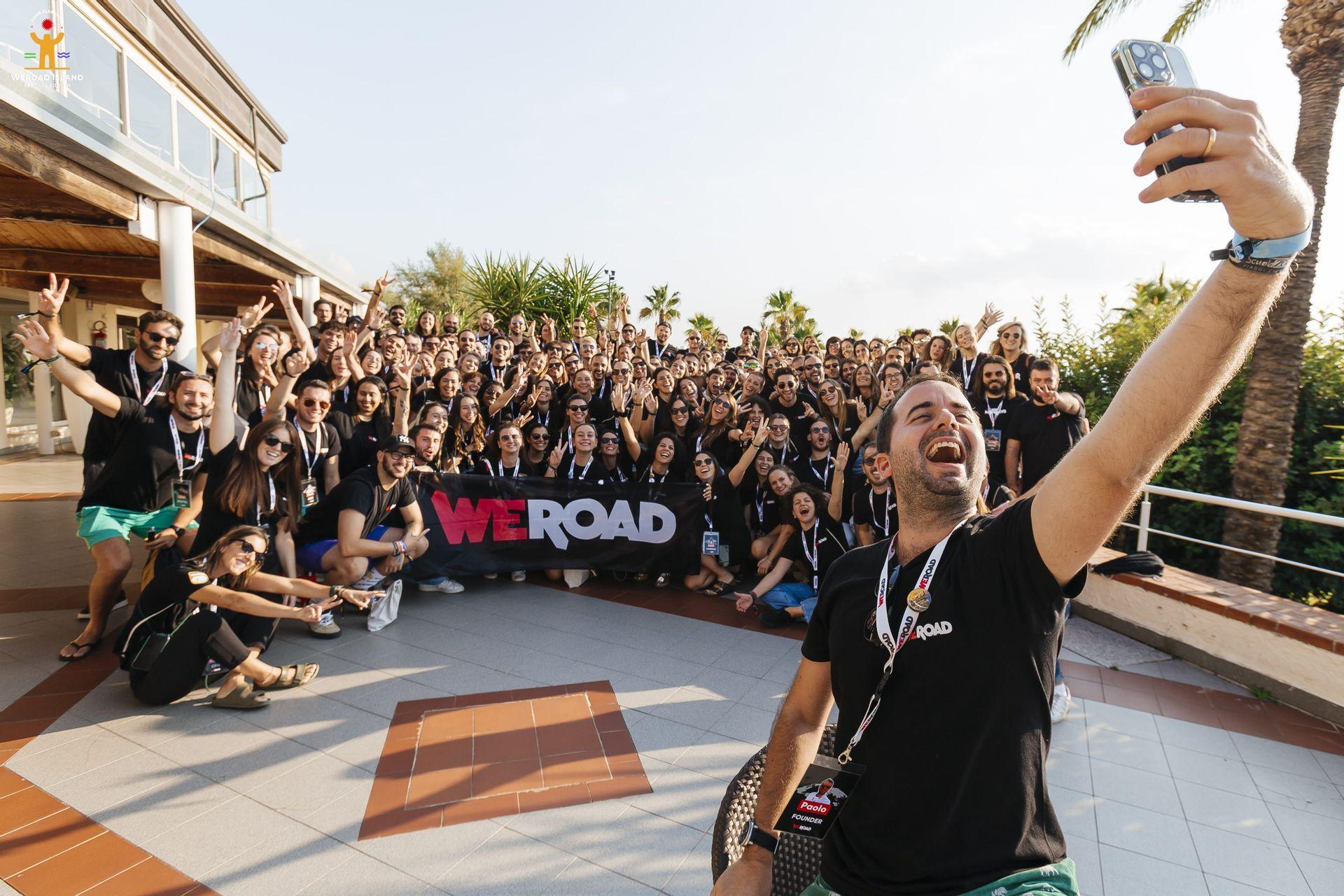Un uomo ride mentre si fa un selfie con un grande gruppo WeRoad in posa con uno striscione su una terrazza soleggiata.