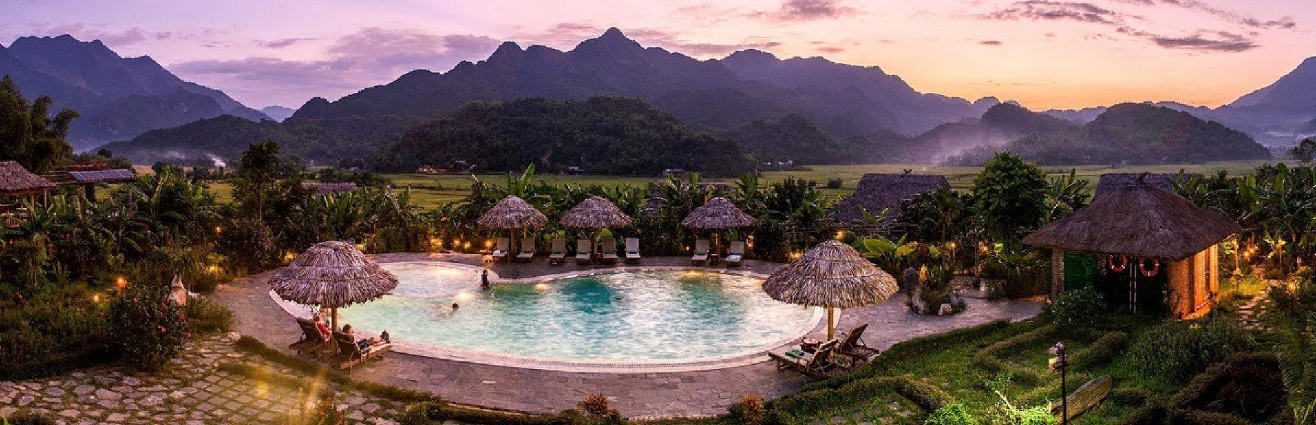 Un edificio tradizionale in legno accanto a una piscina a sfioro, che riflette il cielo e le nuvole al tramonto, con montagne sullo sfondo.