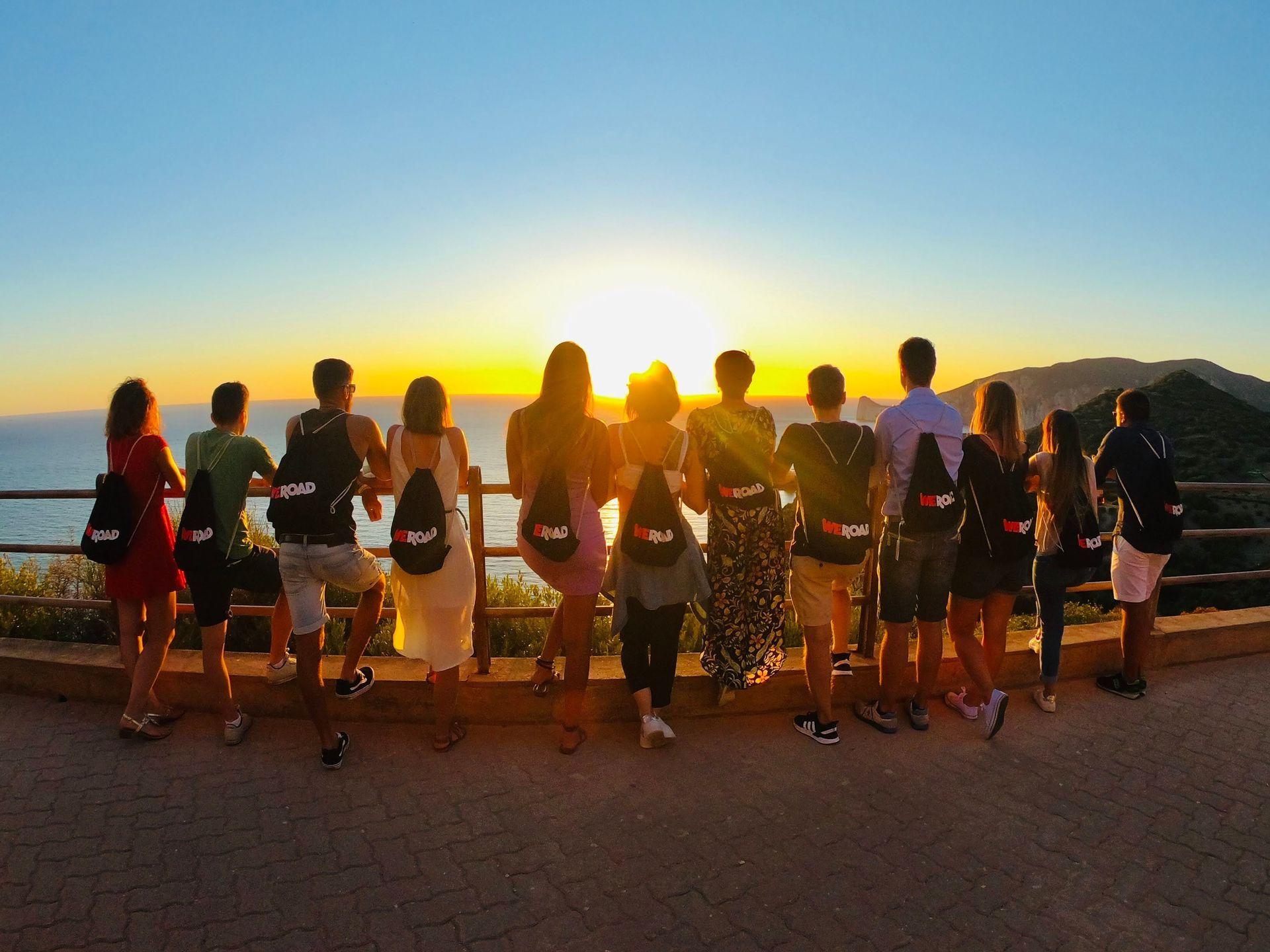 Un gruppo WeRoad, ripreso di spalle, ammira il tramonto sull'oceano da un parapetto.