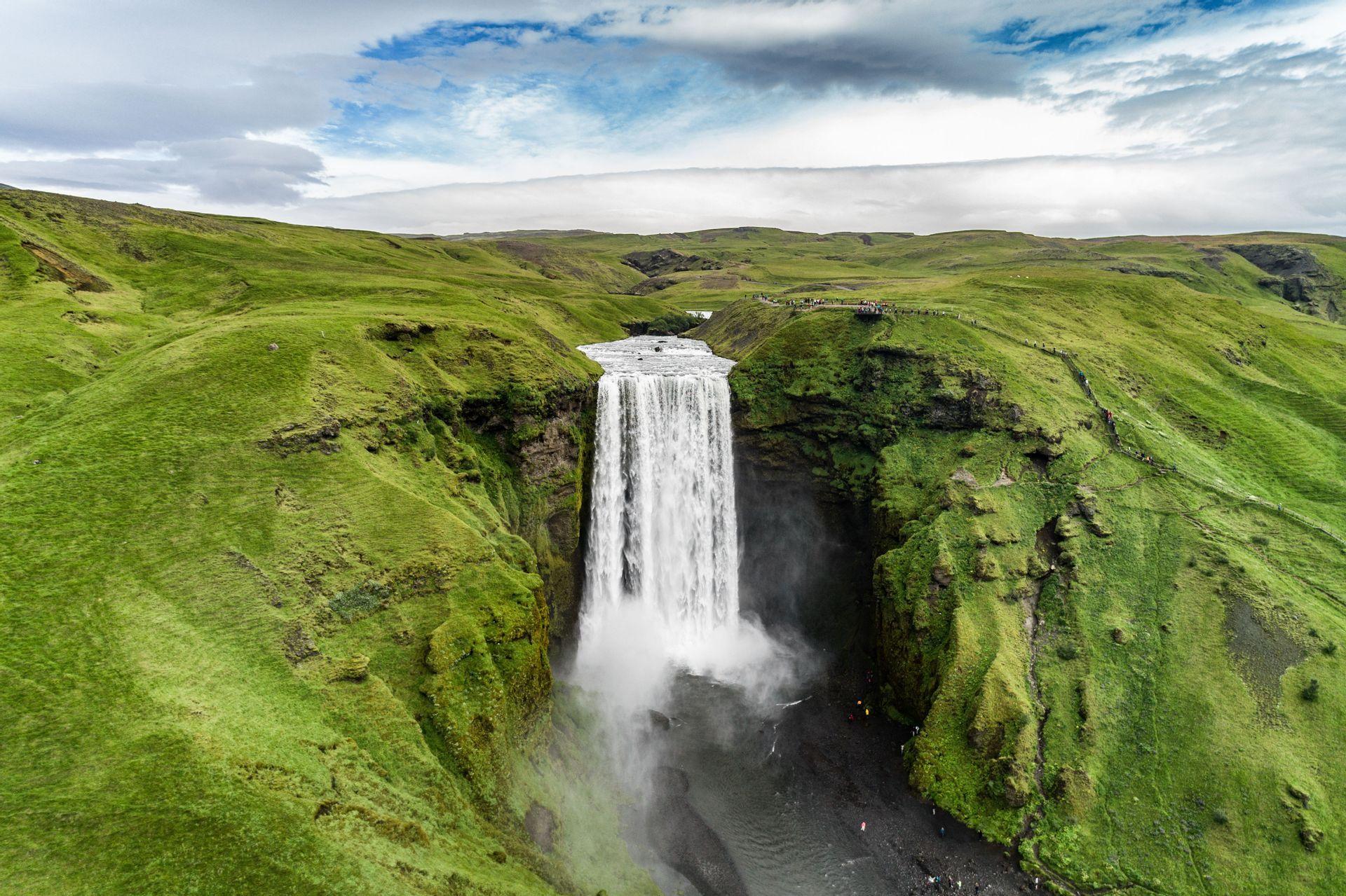 Una vista aérea de una gran cascada que cae entre exuberantes colinas verdes, con un grupo de viaje de WeRoad observando desde arriba y abajo.