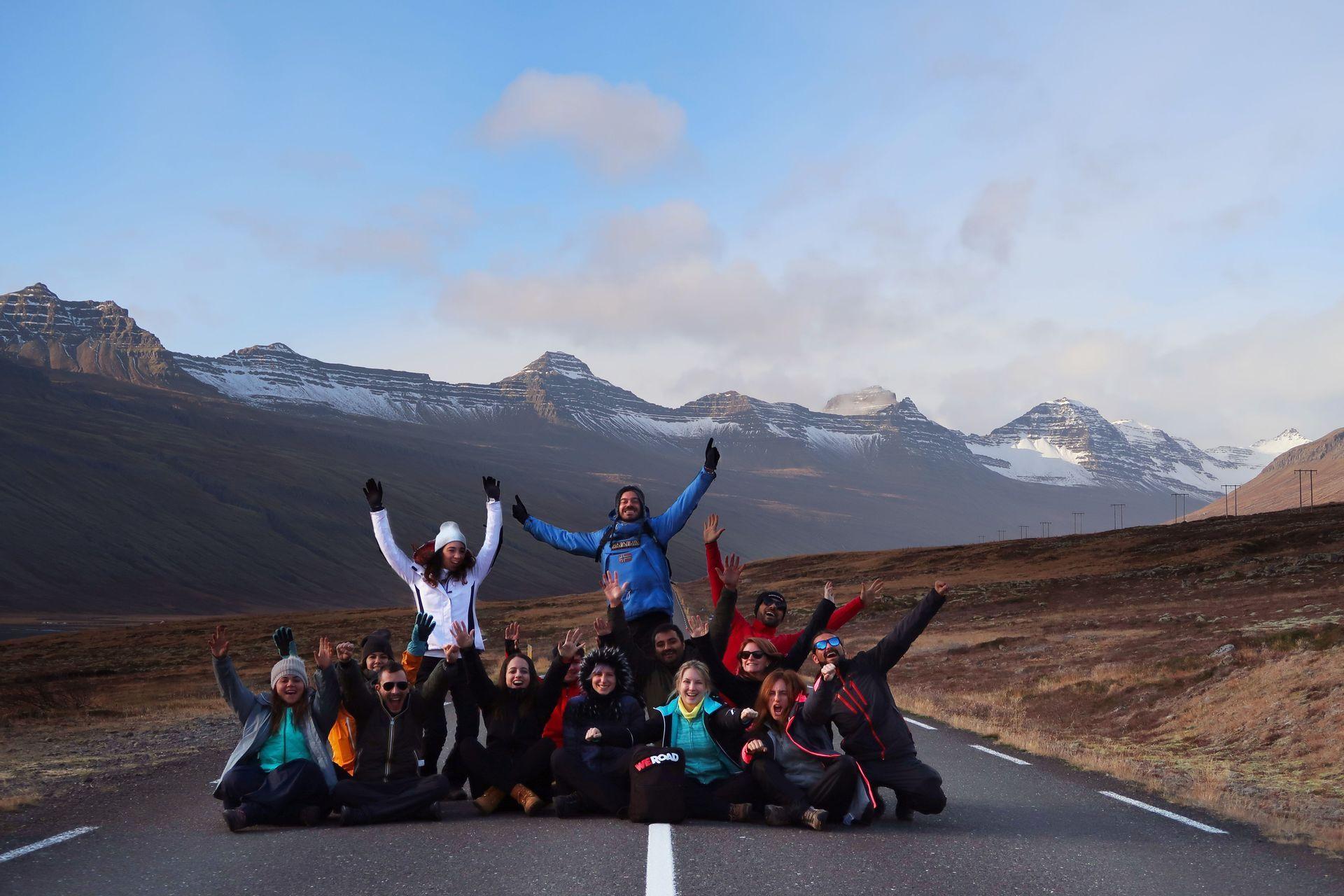 A WeRoad group trip posing on an empty road, with their arms raised in front of a snow-capped mountain range under a clear sky.