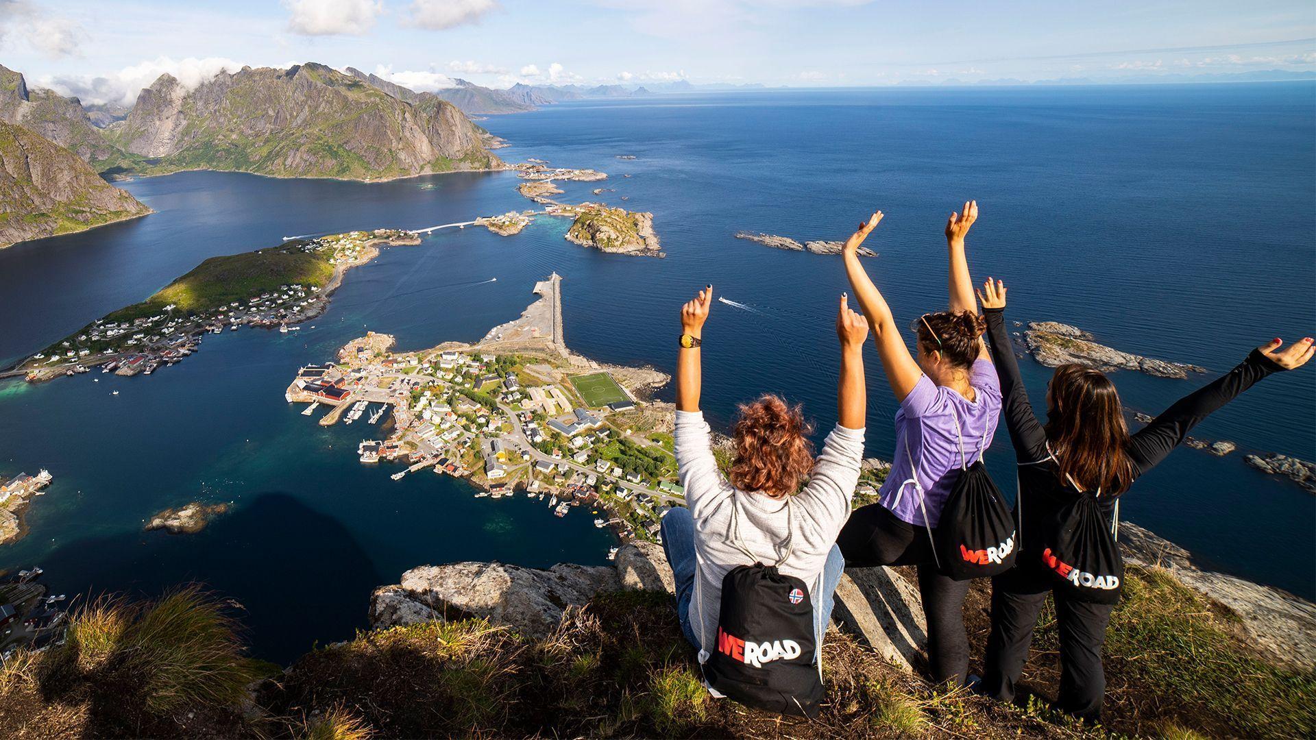 A WeRoad group trip of three women sitting on a clifftop with arms raised, overlooking a coastal village and fjord.