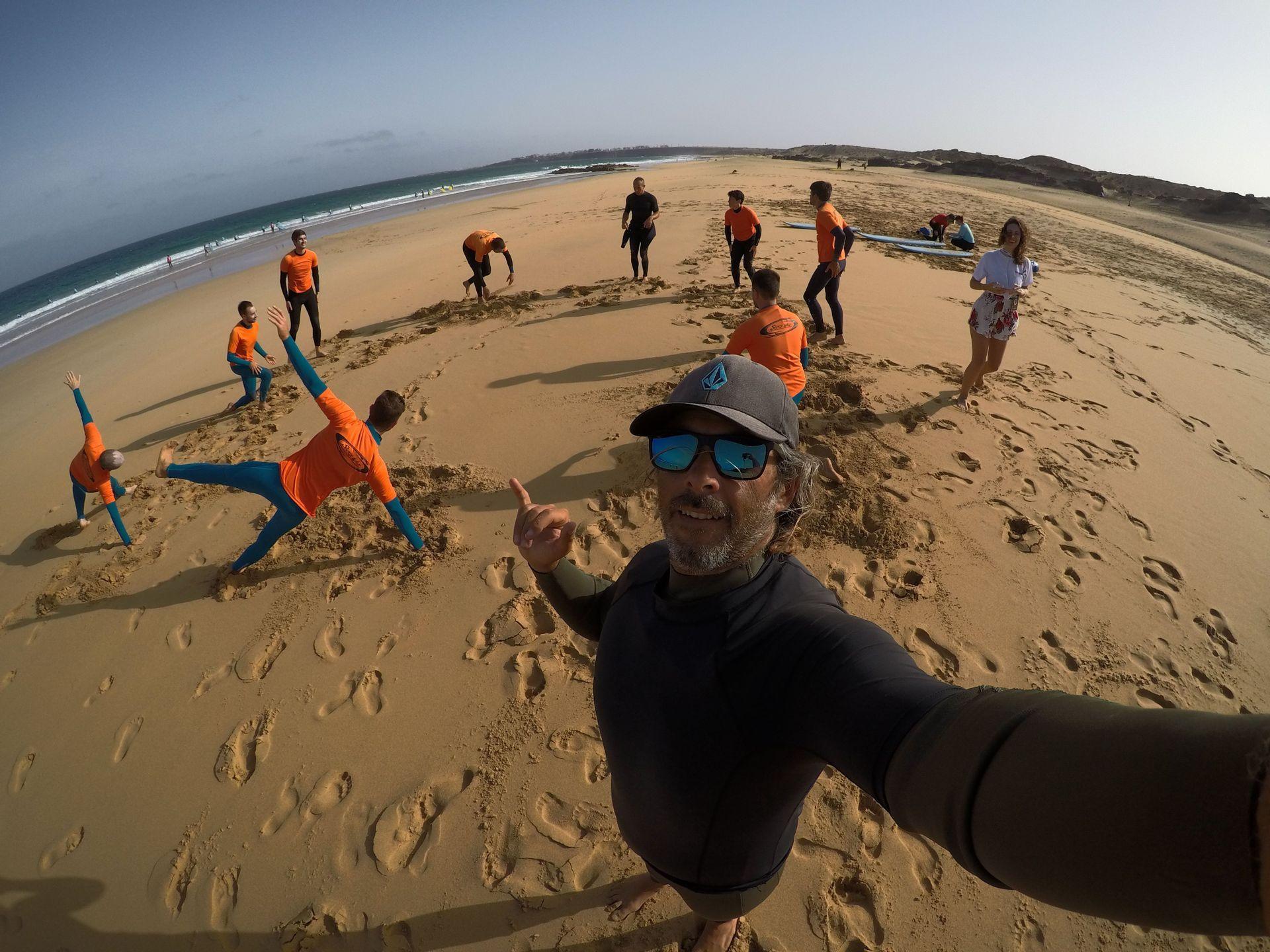 Un uomo si fa un selfie con un gruppo WeRoad mentre si riscalda per una lezione di surf su una spiaggia sabbiosa.