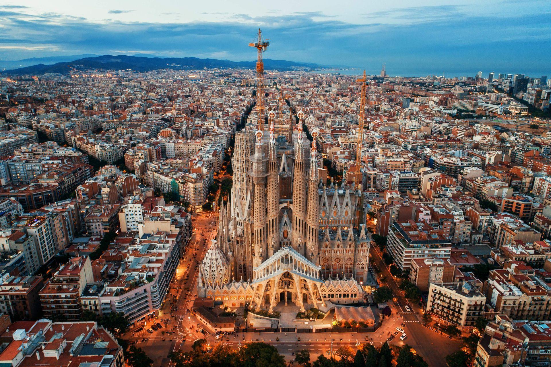 An aerial view of a large, intricate cathedral at dusk, surrounded by a dense cityscape with lights on.