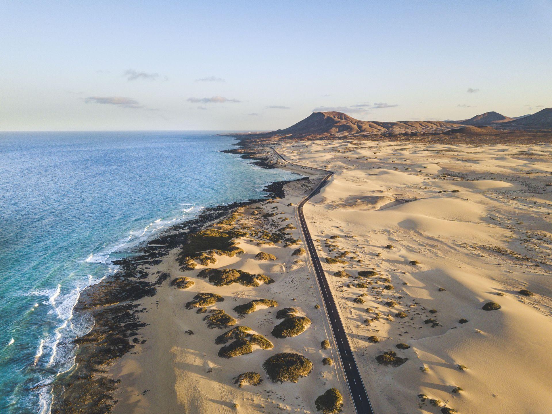 Vista aerea di una strada asfaltata che si snoda lungo la costa, separando vaste dune di sabbia dall'oceano blu.