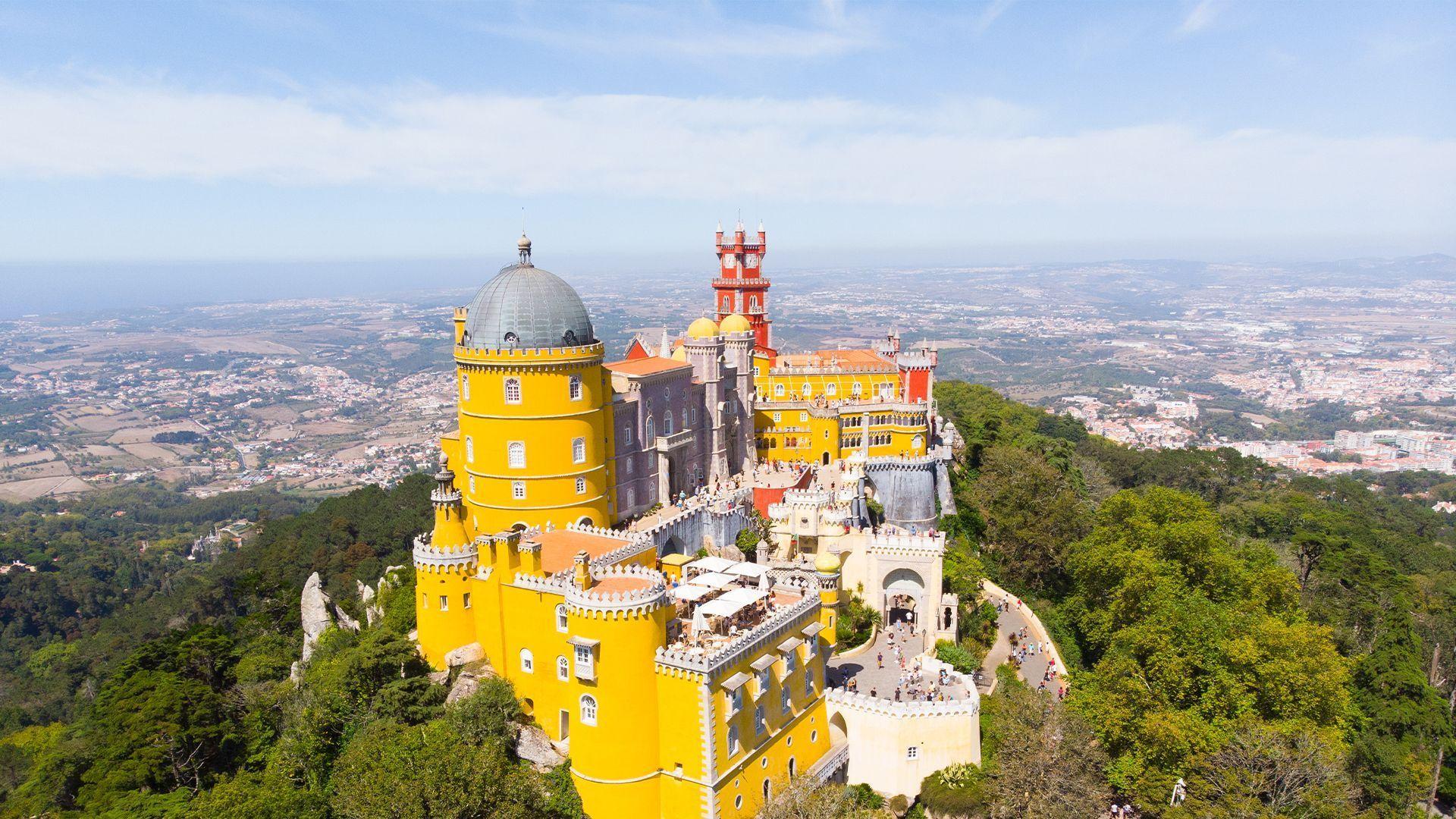 Vue aérienne d'un palais jaune et rouge éclatant sur une colline boisée, avec une vaste ville en arrière-plan.
