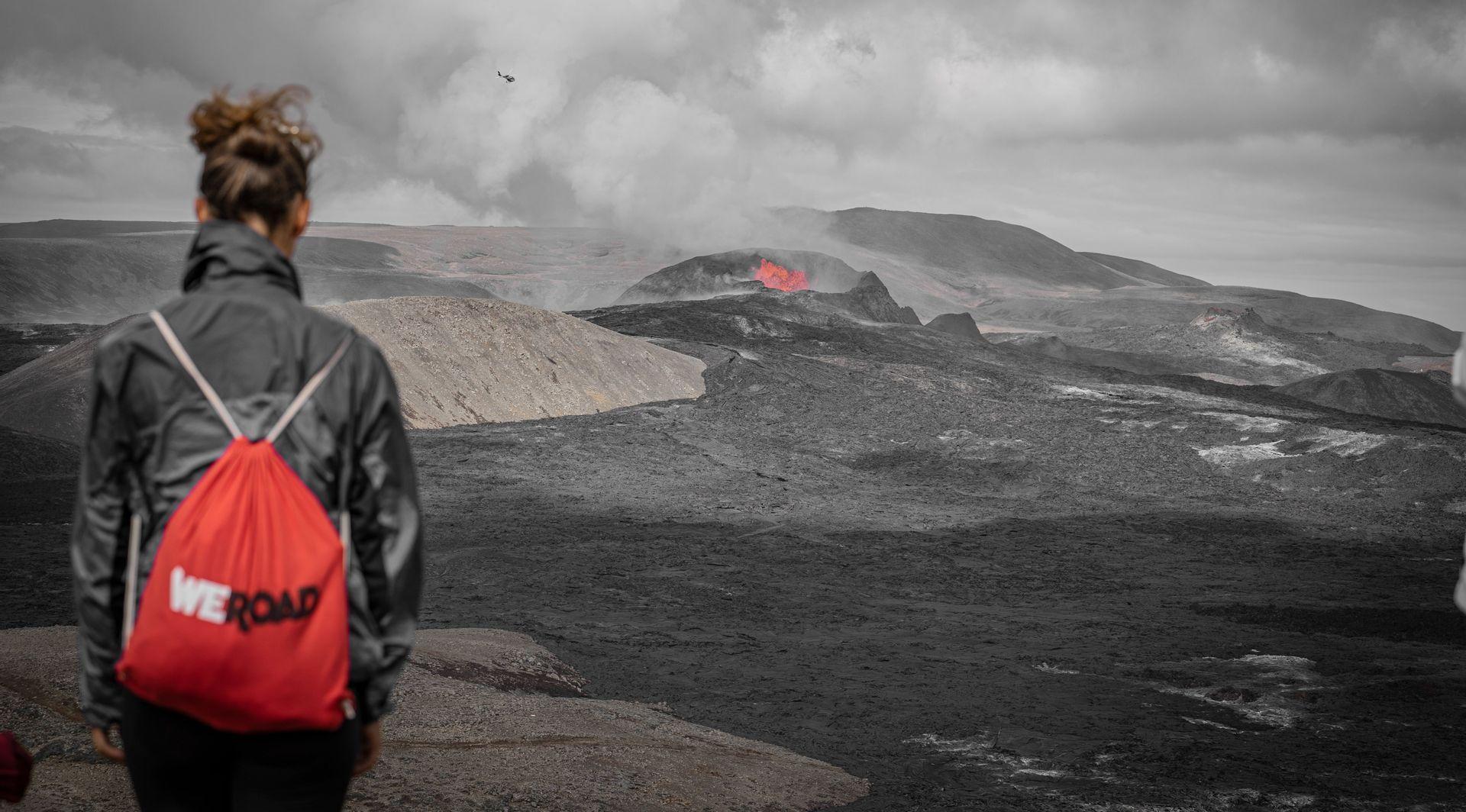 Una persona di spalle con uno zaino rosso WeRoad guarda un vulcano in eruzione su un paesaggio scuro e roccioso.