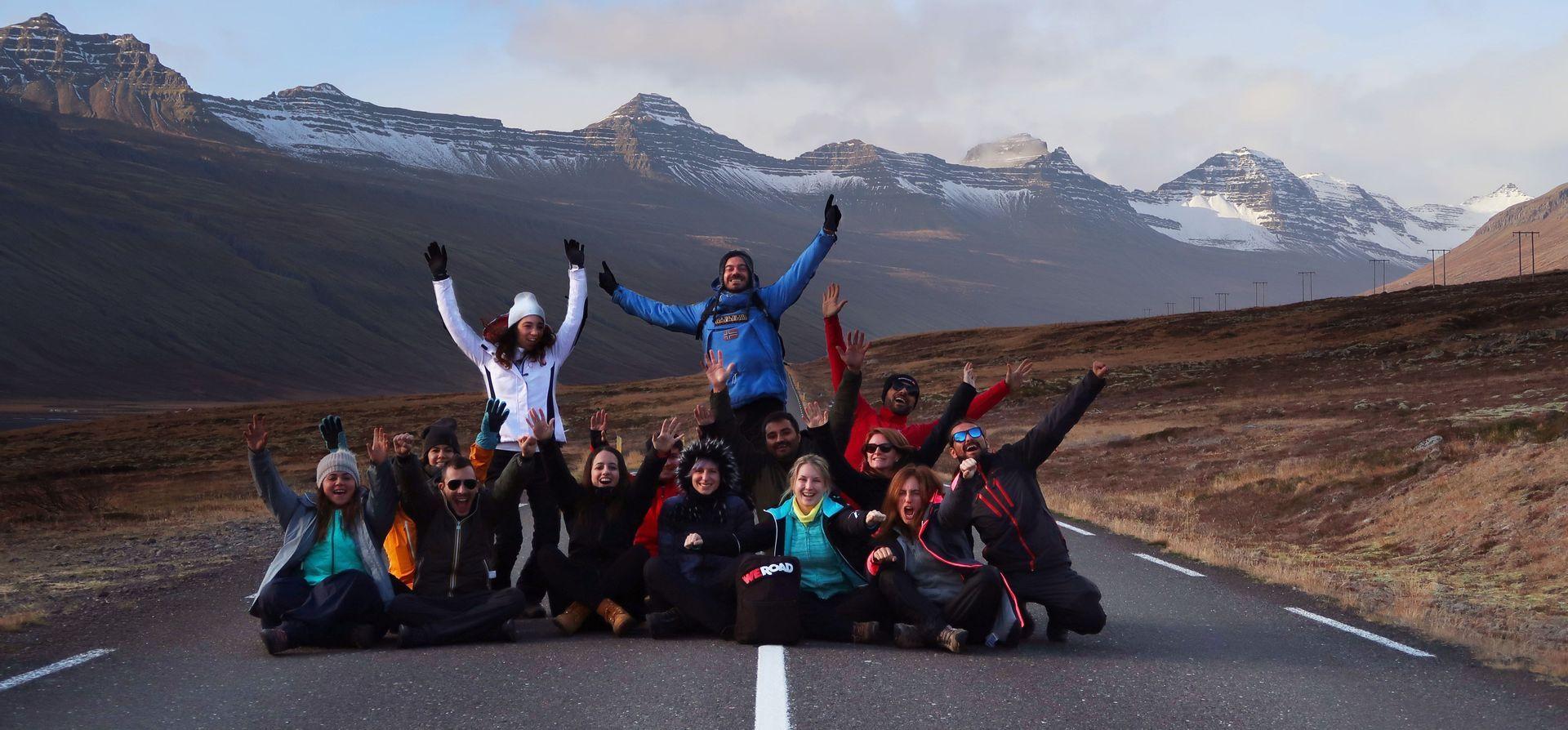 Un gruppo WeRoad posa per una foto, seduti e in piedi su una strada con uno sfondo di montagne innevate.