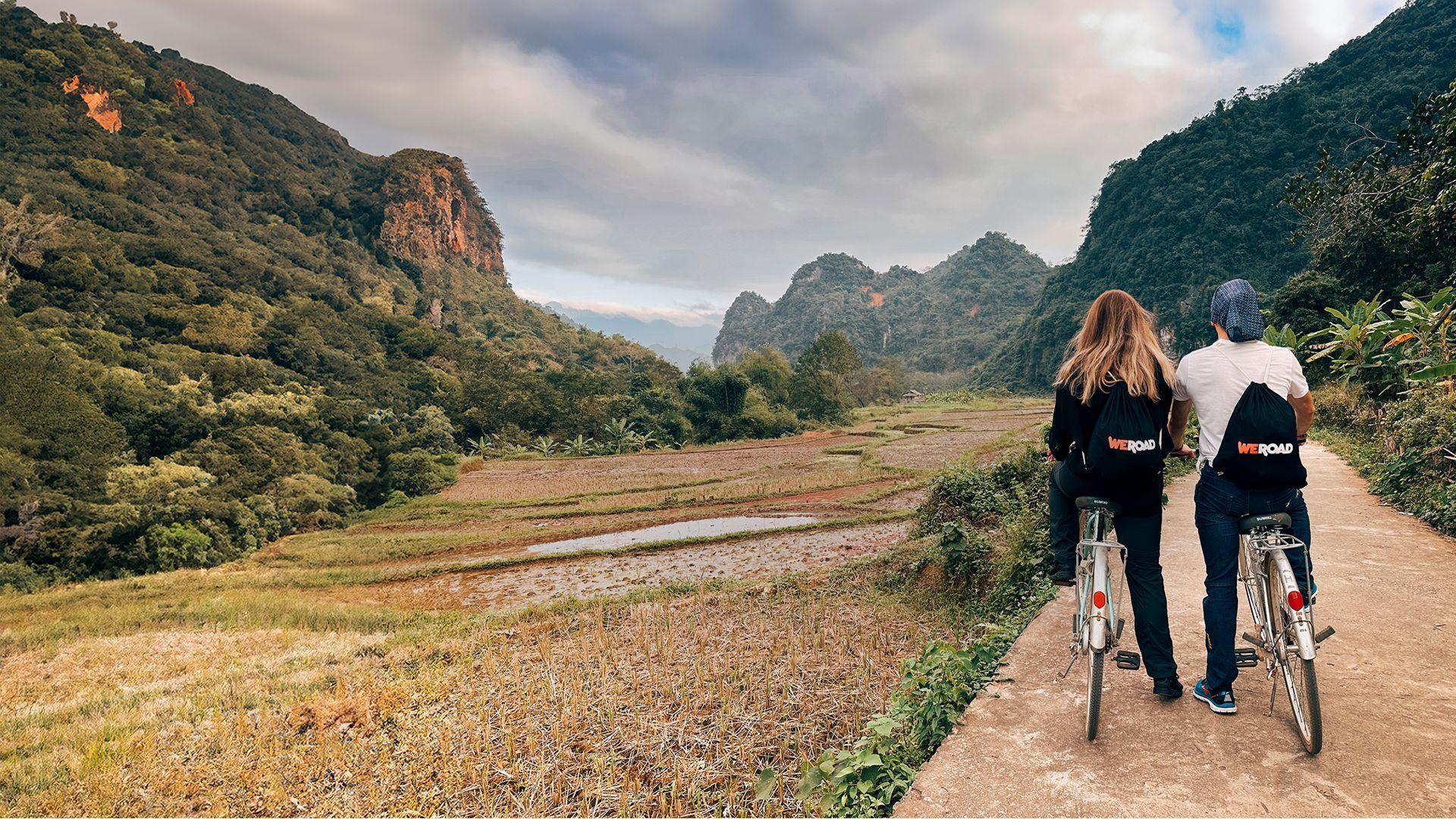 Dos personas de un viaje en grupo de WeRoad andan en bicicleta por un sendero con vistas a campos de arroz en terrazas y montañas verdes y exuberantes.