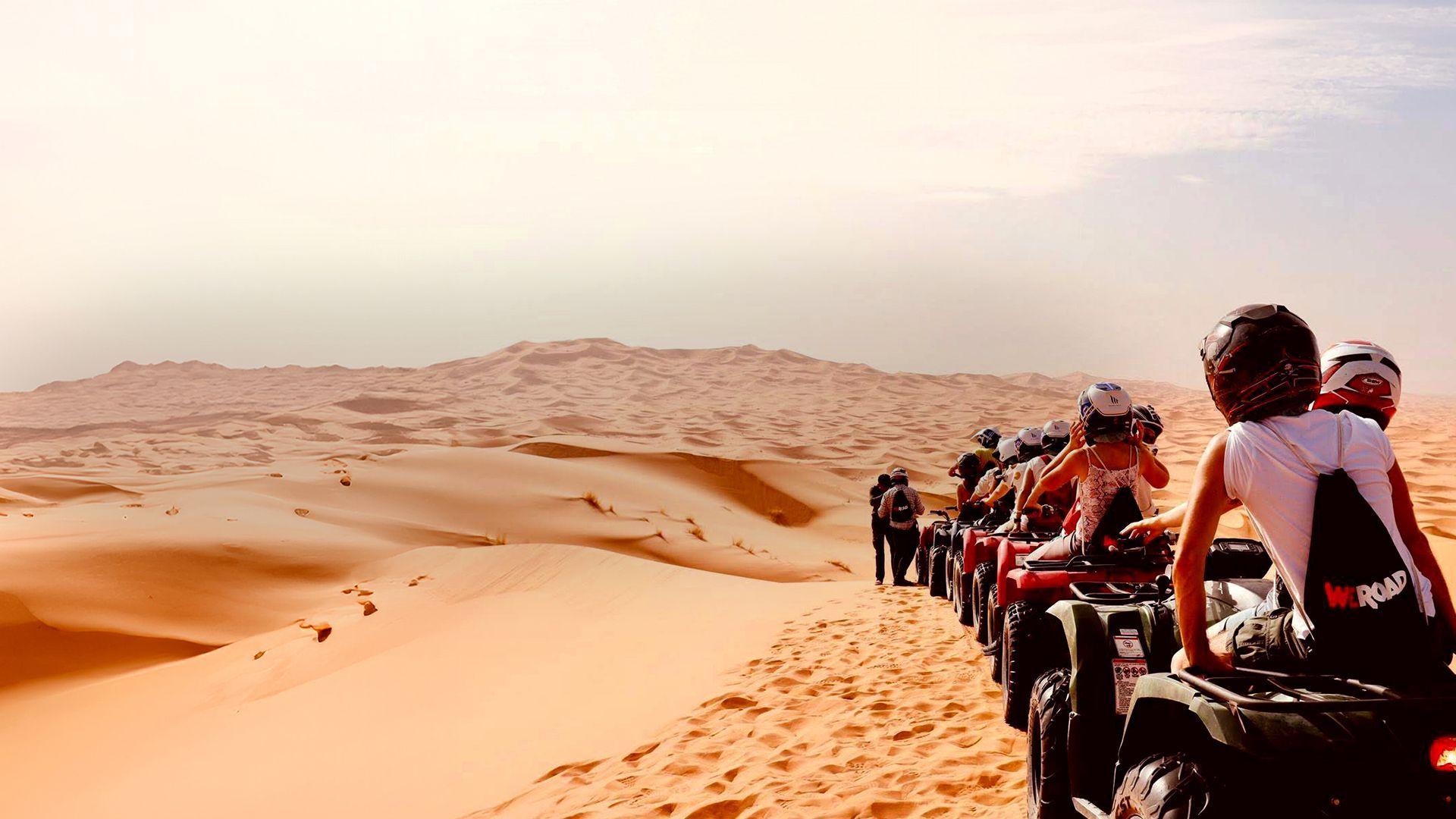 Un gruppo WeRoad con caschi in sella a quad allineati sulle dune di sabbia del deserto sotto un cielo luminoso.