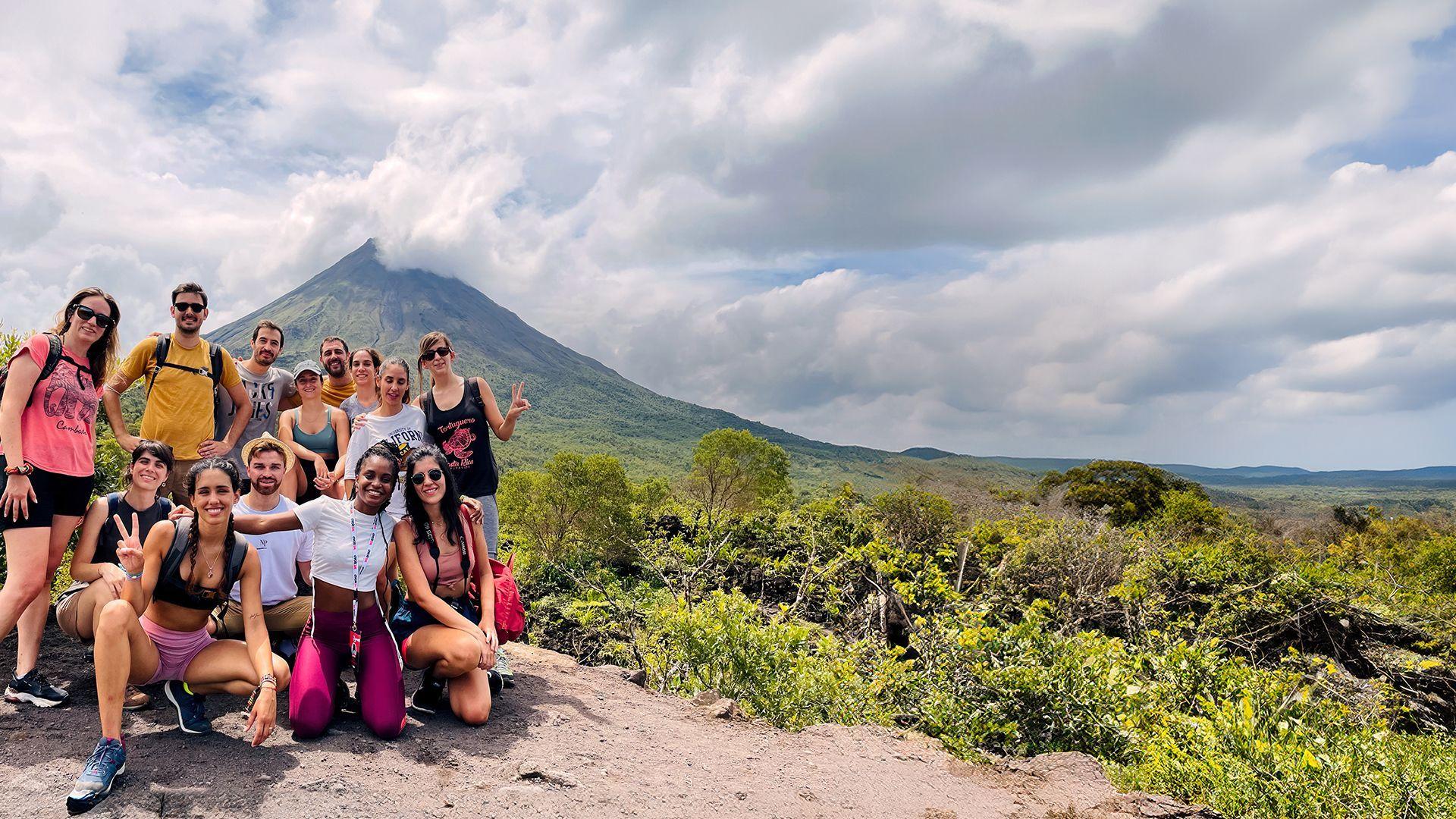 Un viaje en grupo de WeRoad posando para una foto en un mirador rocoso con un gran volcán cubierto de nubes de fondo.