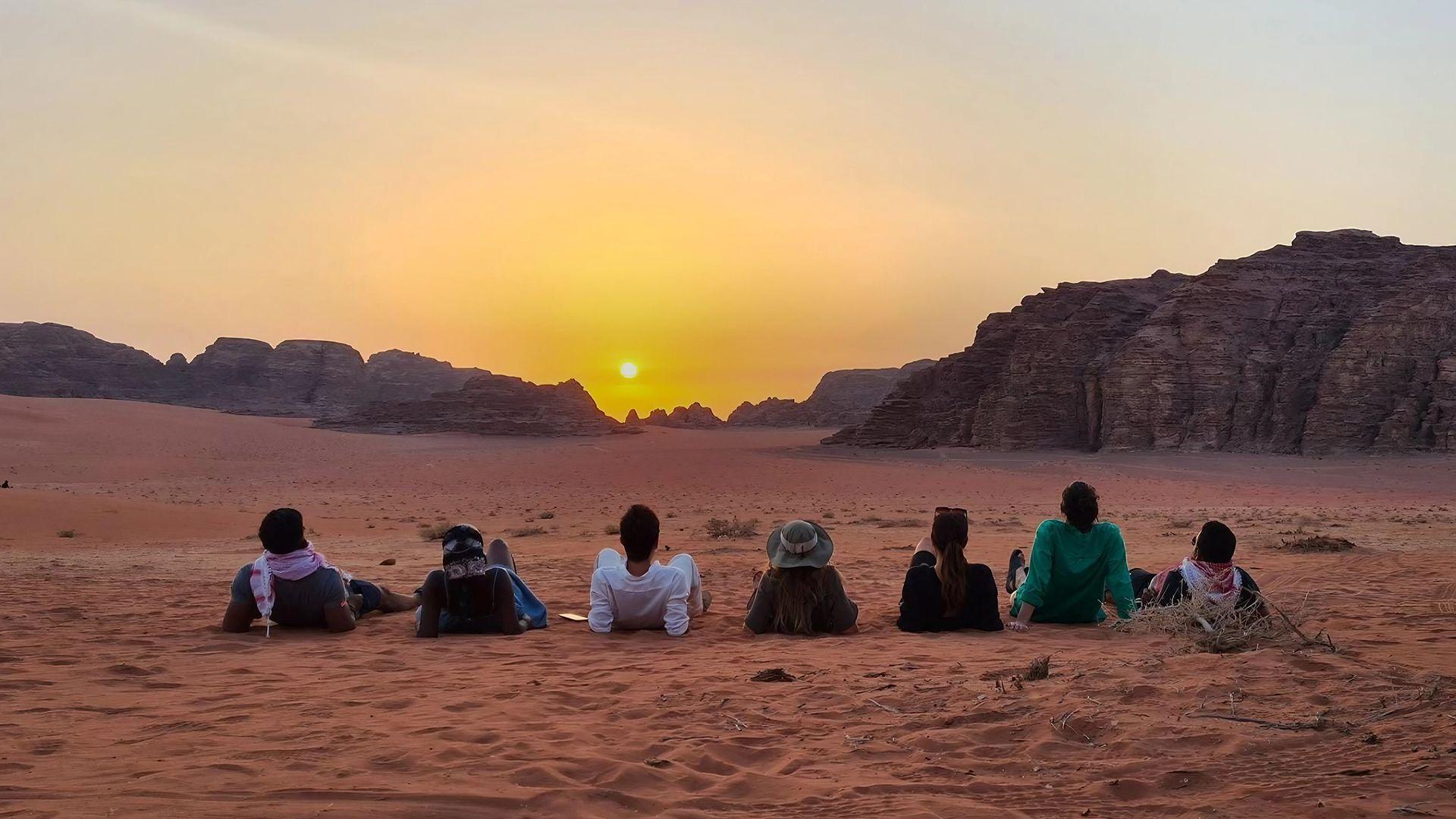 Un viaje en grupo de WeRoad se sienta en la arena roja del desierto viendo el atardecer detrás de montañas rocosas distantes.