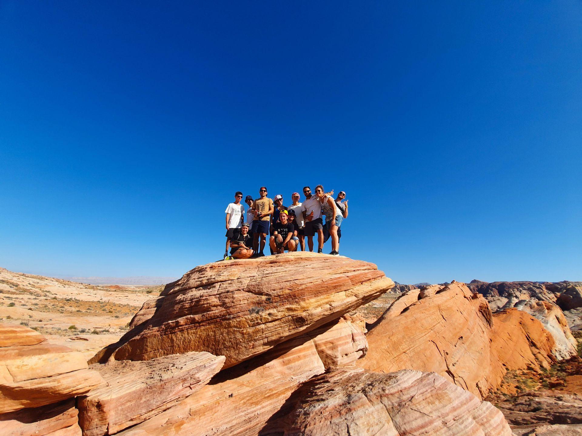 Un grupo de viaje de WeRoad posando juntos en una gran formación rocosa roja en un paisaje desértico bajo un cielo azul claro.