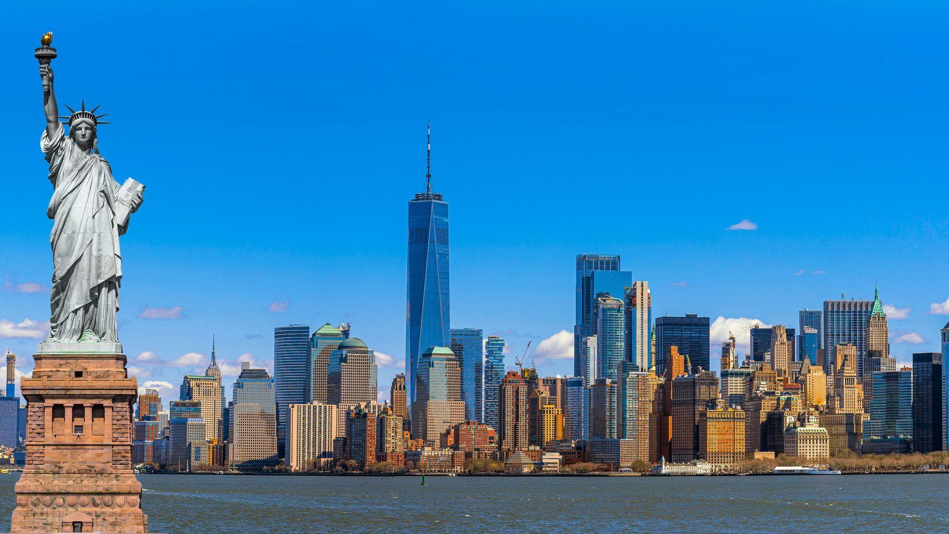 La Statua della Libertà si erge maestosa sul suo piedistallo, con lo skyline di Manhattan visibile attraverso l'acqua sotto un cielo azzurro e sereno.