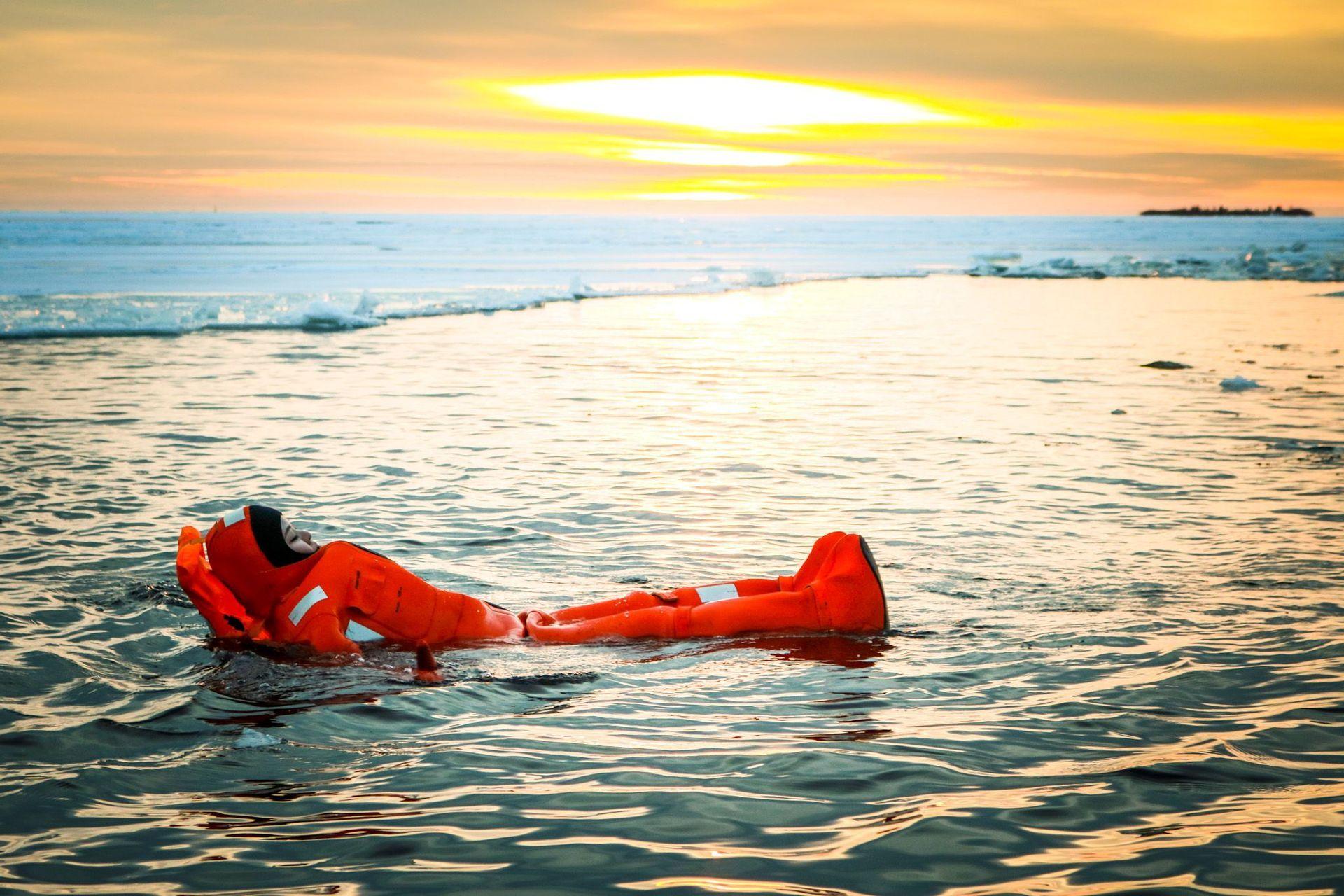 Une personne en combinaison de survie rouge flotte sur le dos dans l'eau glacée, créant des ondulations près d'un rivage gelé au coucher du soleil.