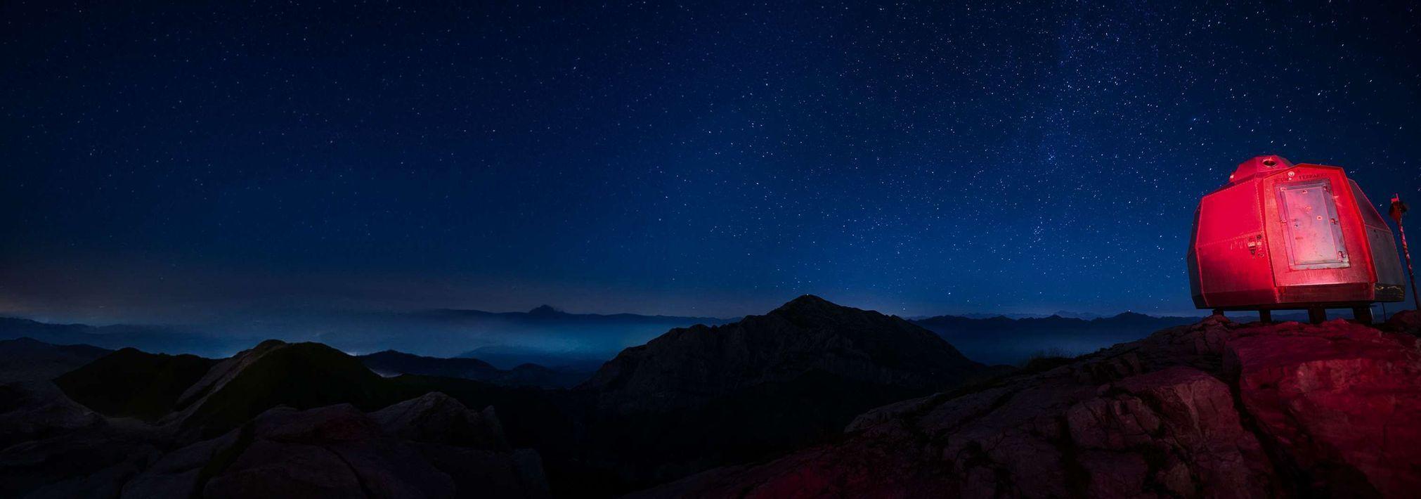 Una cabaña roja e iluminada se encuentra en una cresta montañosa rocosa bajo un cielo estrellado.