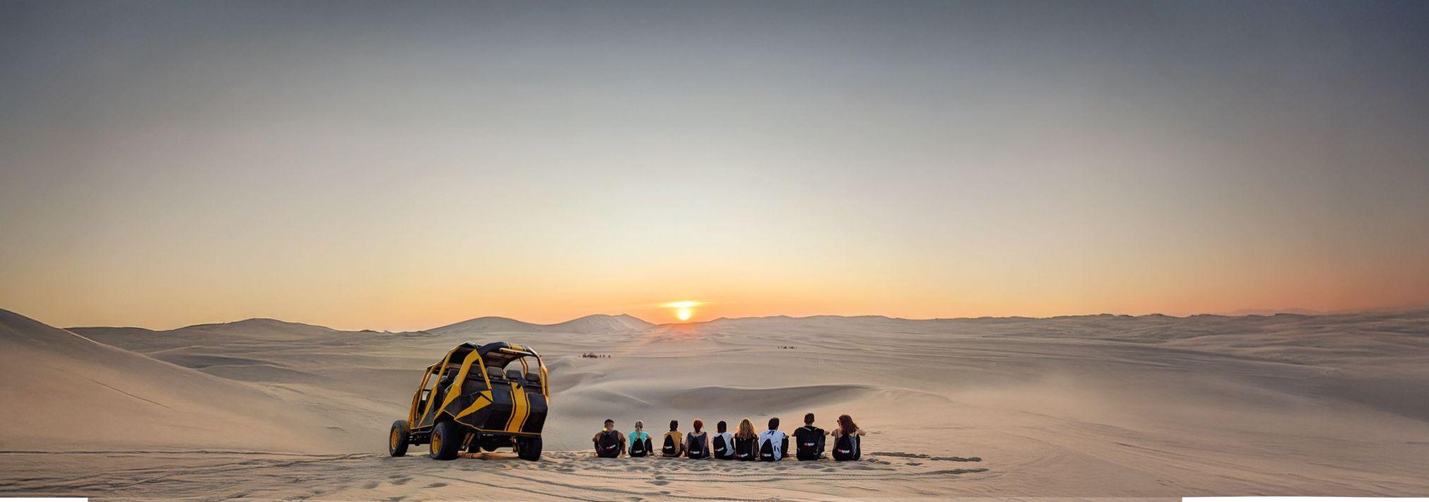 A WeRoad group trip sits on a sand dune watching the sunset, with a yellow buggy parked in the desert landscape.