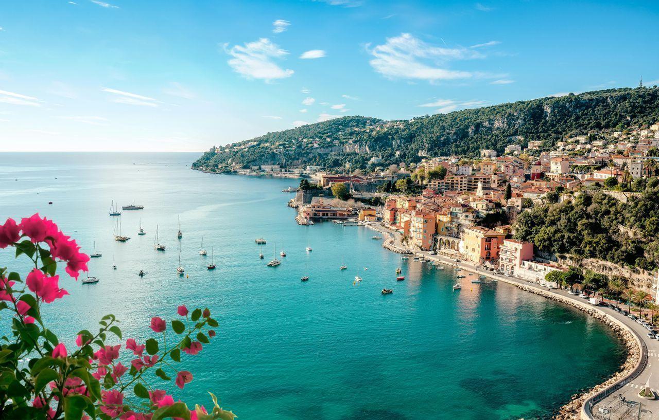 Une ville côtière colorée à flanc de colline domine une baie turquoise avec de nombreux voiliers et des fleurs roses au premier plan.