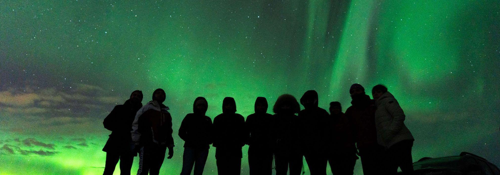 Siluetas de un viaje en grupo WeRoad bajo la aurora boreal verde y un cielo nocturno estrellado.