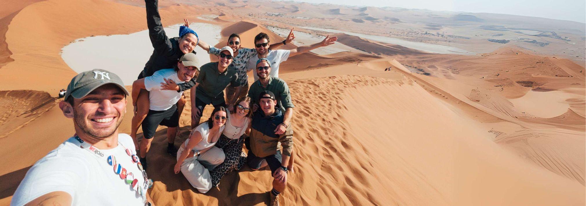 Un voyage de groupe WeRoad sourit et pose pour un selfie sur une grande dune de sable dans le désert.