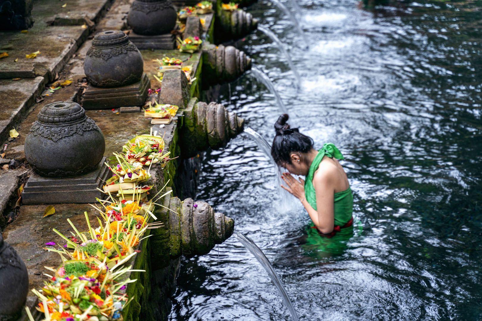 Una donna con un top verde esegue un rito di purificazione, chinando la testa sotto un getto d'acqua da una fontana di pietra.