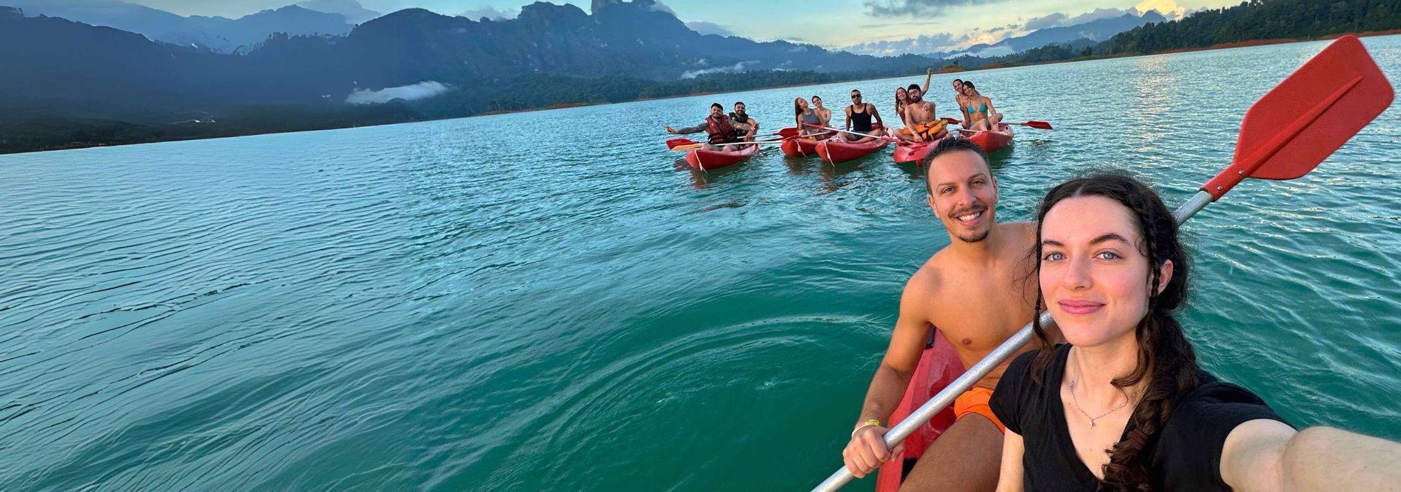 Viaggio di gruppo WeRoad in kayak su un lago, con una coppia in primo piano che si scatta un selfie nel loro kayak rosso.