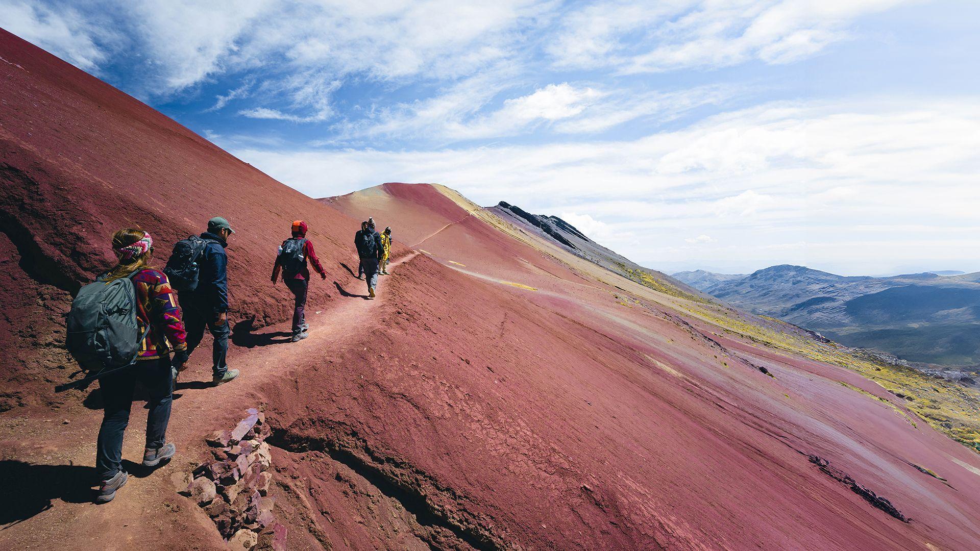 Un viaje en grupo de WeRoad con mochilas haciendo senderismo por un sendero estrecho en una montaña roja bajo un cielo parcialmente nublado.