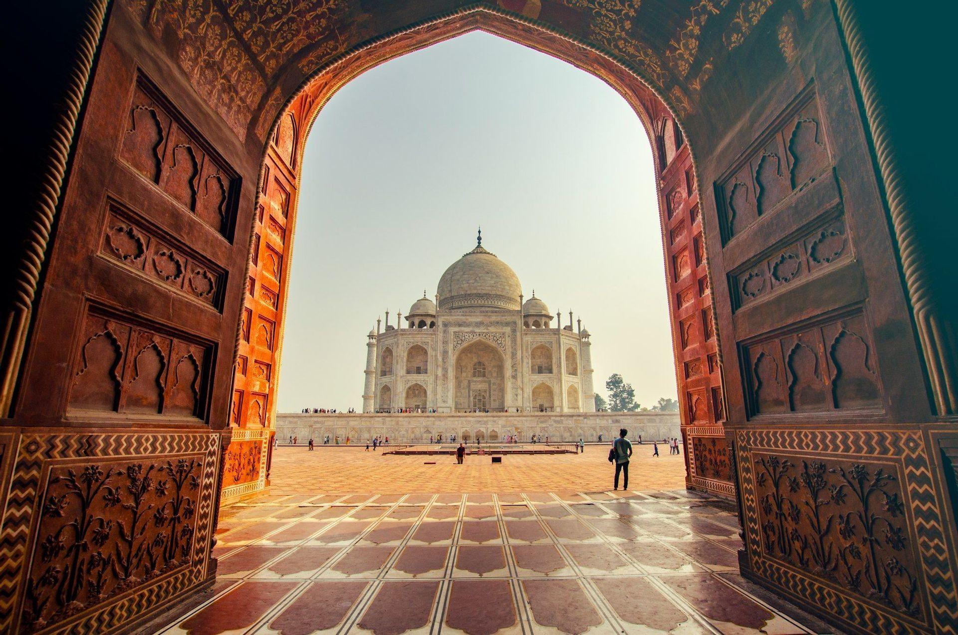 Una vista del Taj Mahal enmarcado por un arco ornamentado de arenisca roja, con gente caminando por la plaza a lo lejos.