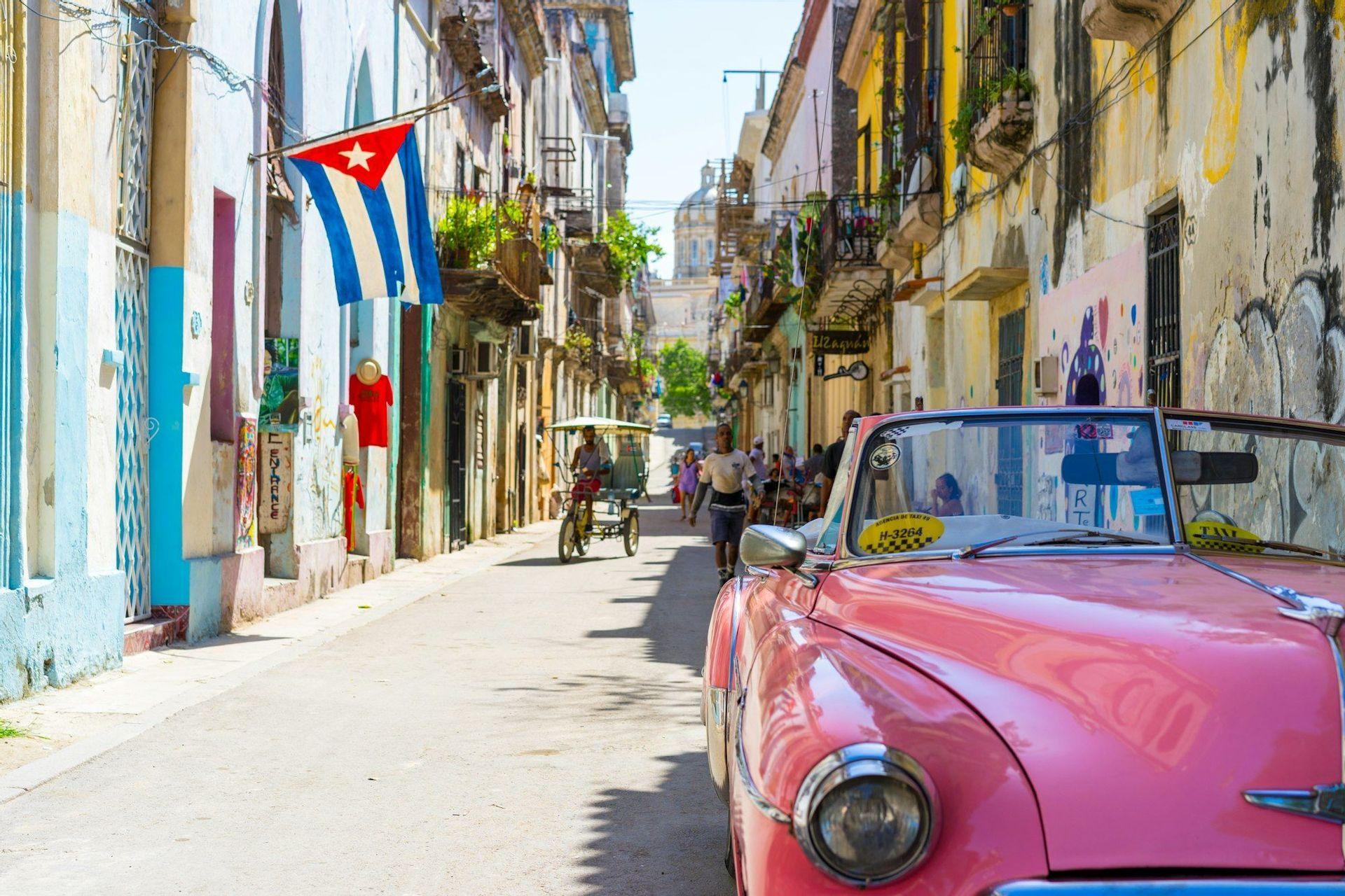 Une voiture décapotable classique rose est garée dans une rue ensoleillée de la ville bordée de bâtiments colorés, avec un drapeau cubain accroché à l'un d'eux.