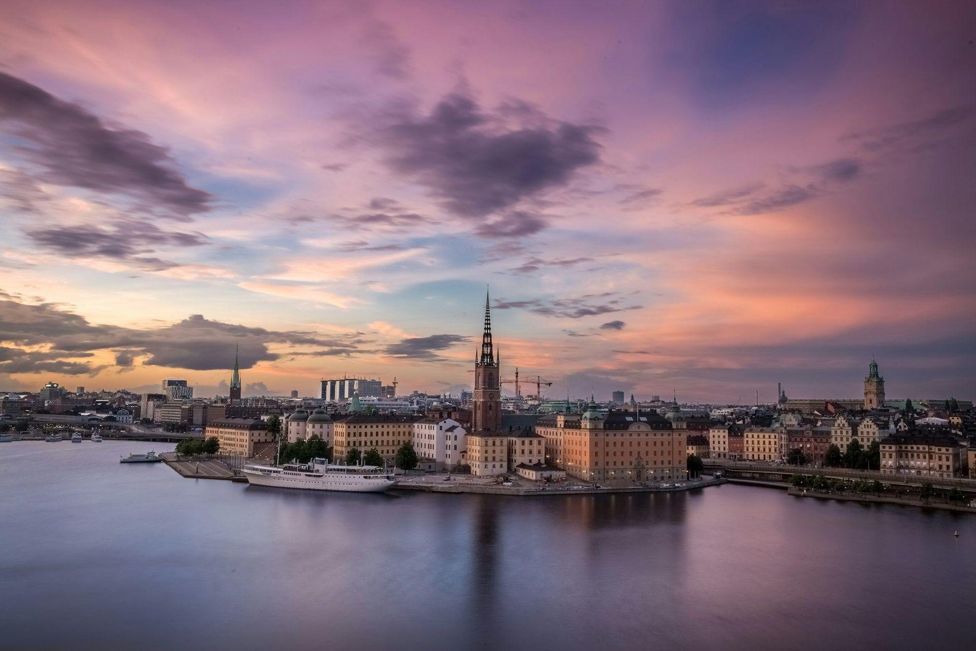 An aerial view of a European cityscape with a prominent church steeple on the waterfront under a colorful sunset sky.