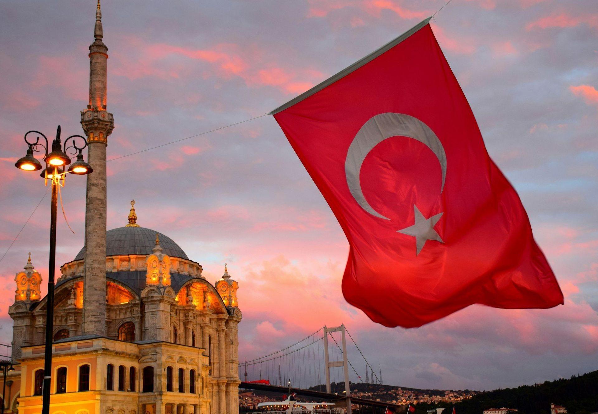 The Turkish flag waves in front of an illuminated mosque and a large suspension bridge against a colorful sunset sky.