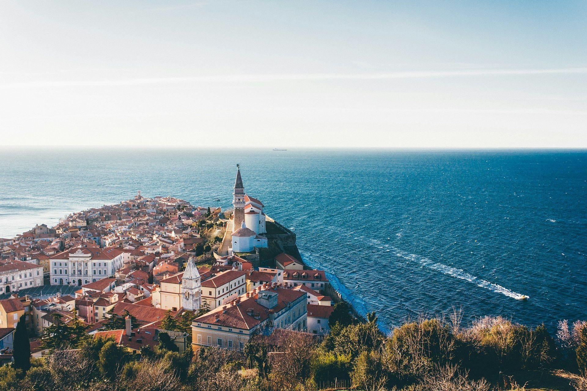Vista panoramica di un borgo costiero con tetti rossi e una chiesa, situato su una penisola che si affaccia sul profondo mare blu.
