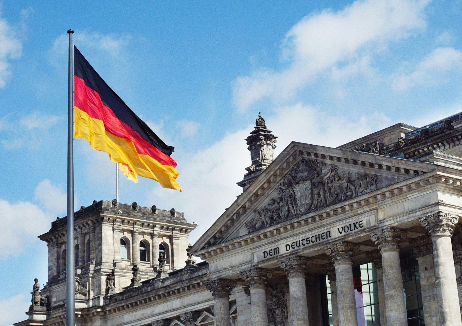 Die deutsche Flagge weht vor der klassischen Fassade des Reichstagsgebäudes unter einem blauen Himmel mit einigen Wolken.