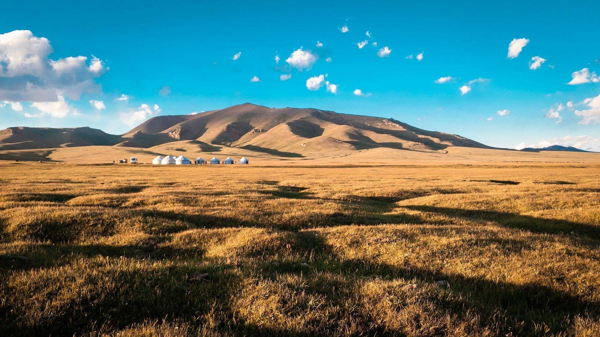 Eine Reihe weißer Jurten steht auf einer weiten, grasbewachsenen Steppe am Fuße eines großen Berges unter einem klaren blauen Himmel.