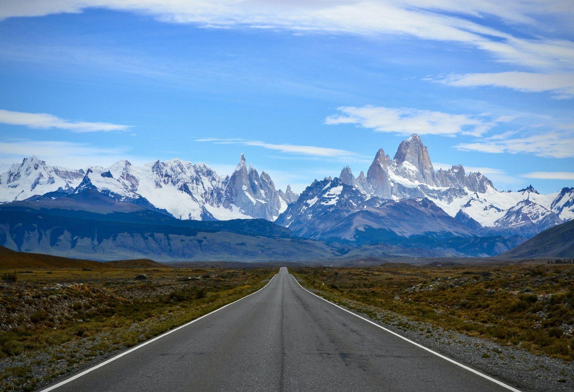 A straight, paved road stretches across a flat, grassy plain towards a distant, snow-capped mountain range under a blue sky.