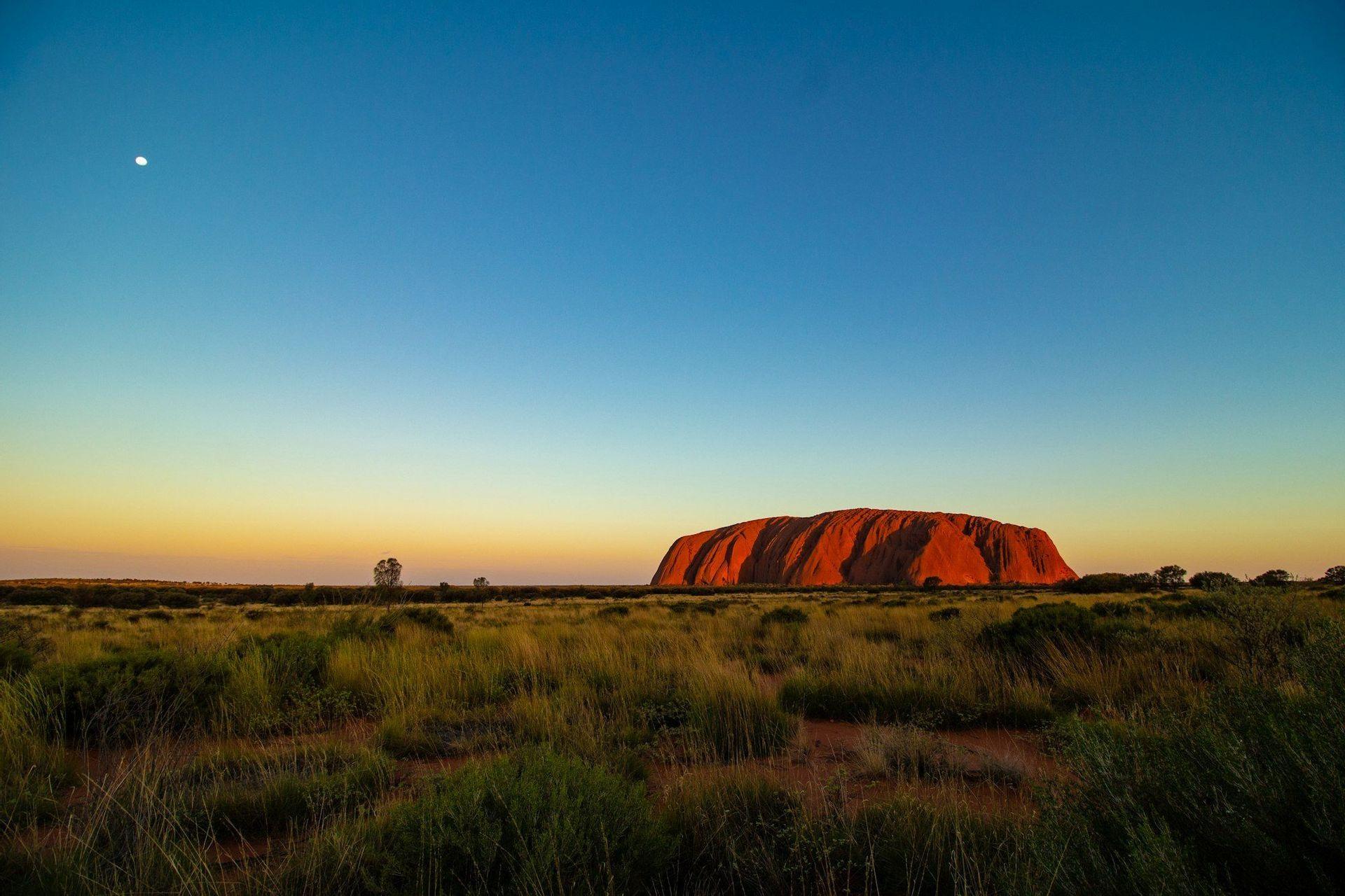 A large red sandstone monolith glows at sunset, rising from a grassy plain under a clear blue sky with the moon visible.