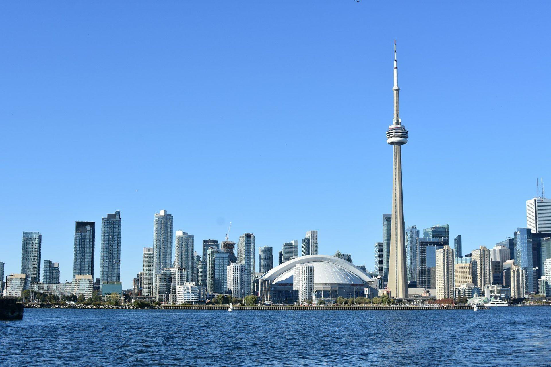 A city skyline with a tall tower and a domed stadium viewed from across the water under a clear blue sky.