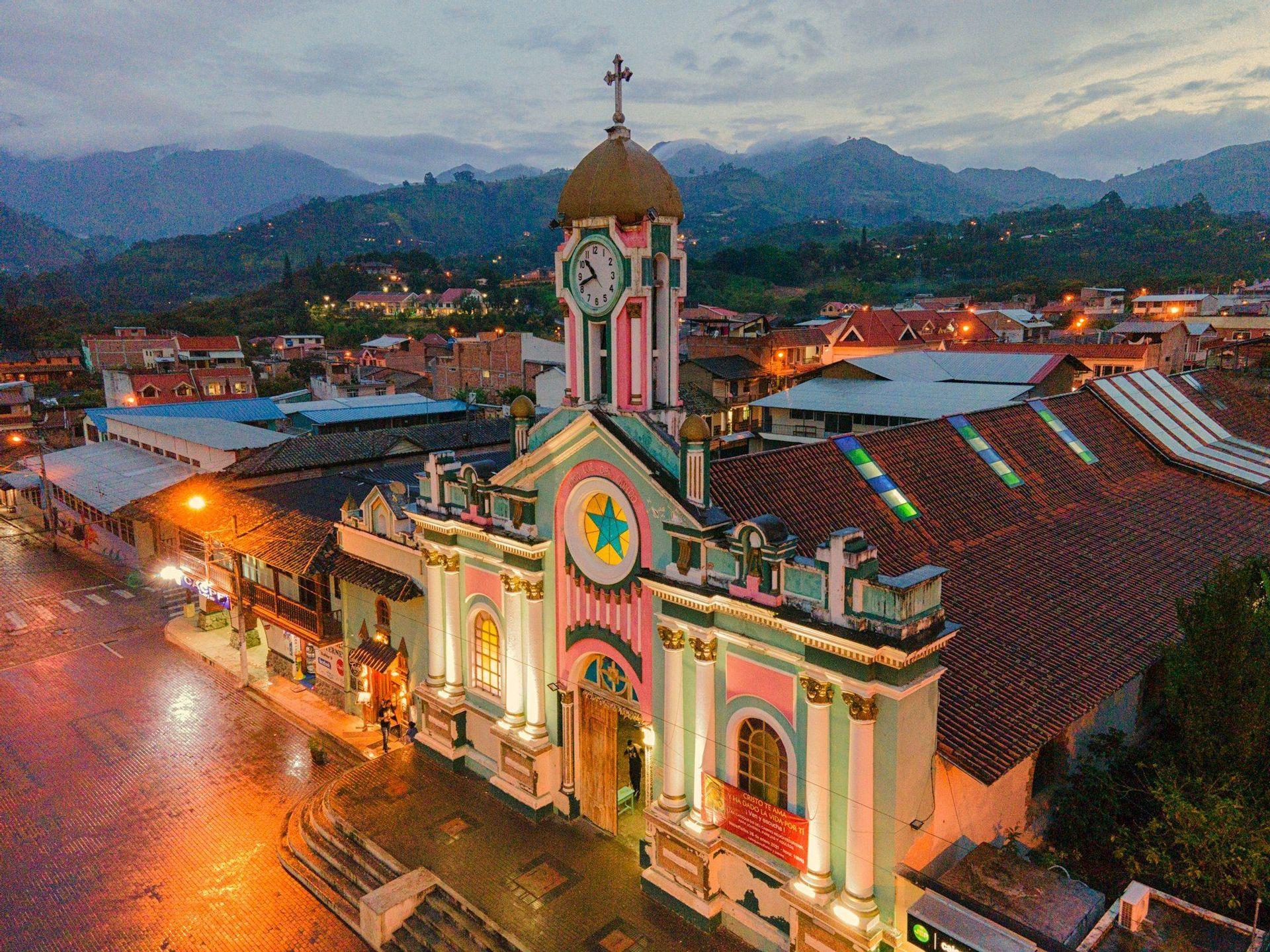 An aerial view of a colorful pink and green church with a clock tower, illuminated at dusk in a small town surrounded by mountains.