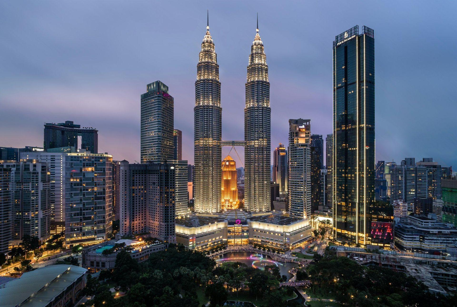 A city skyline with illuminated twin skyscrapers towering over modern buildings and a green park at dusk.