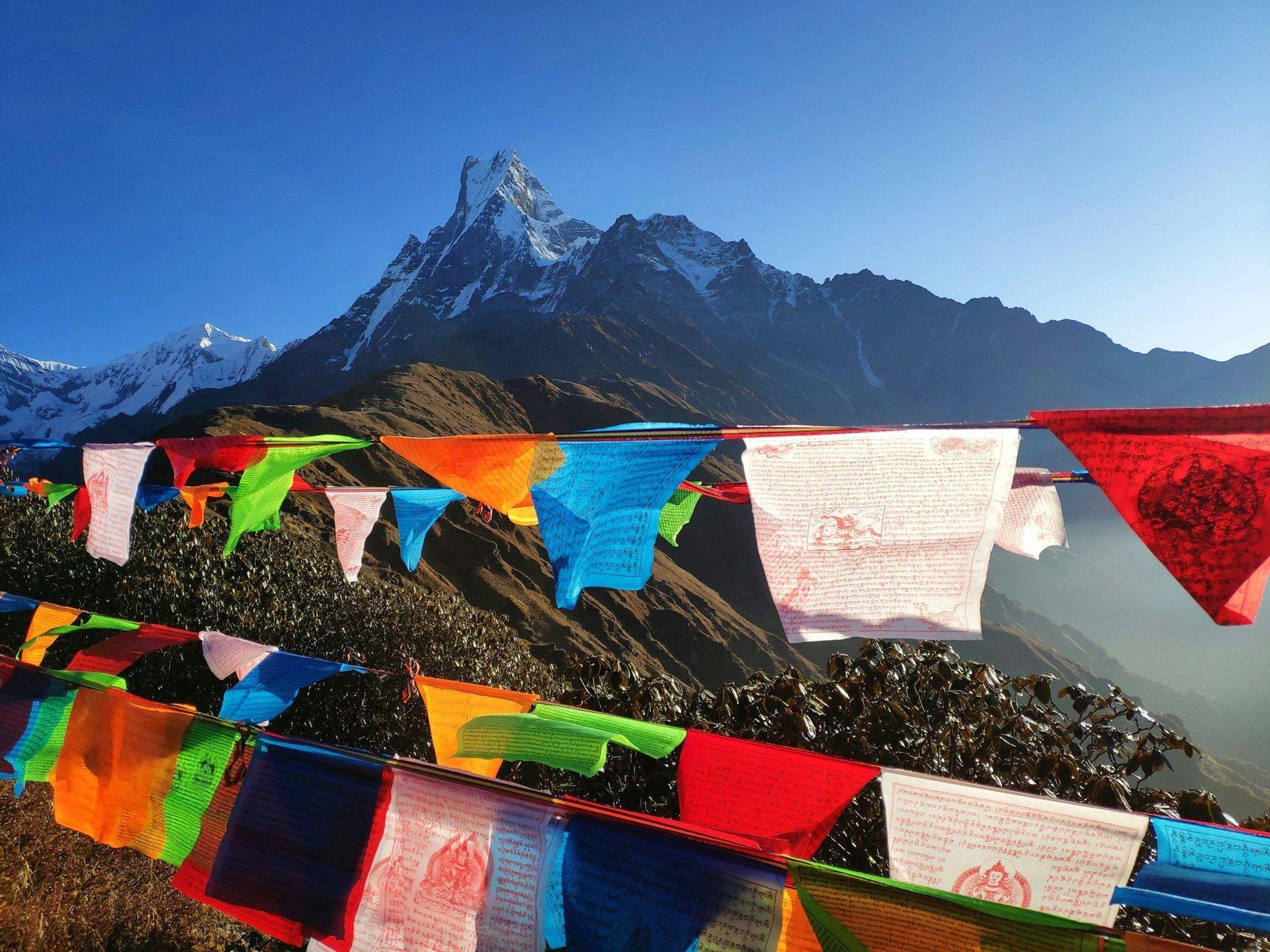 Colorful prayer flags hang in front of a jagged, snow-capped mountain peak against a clear blue sky.