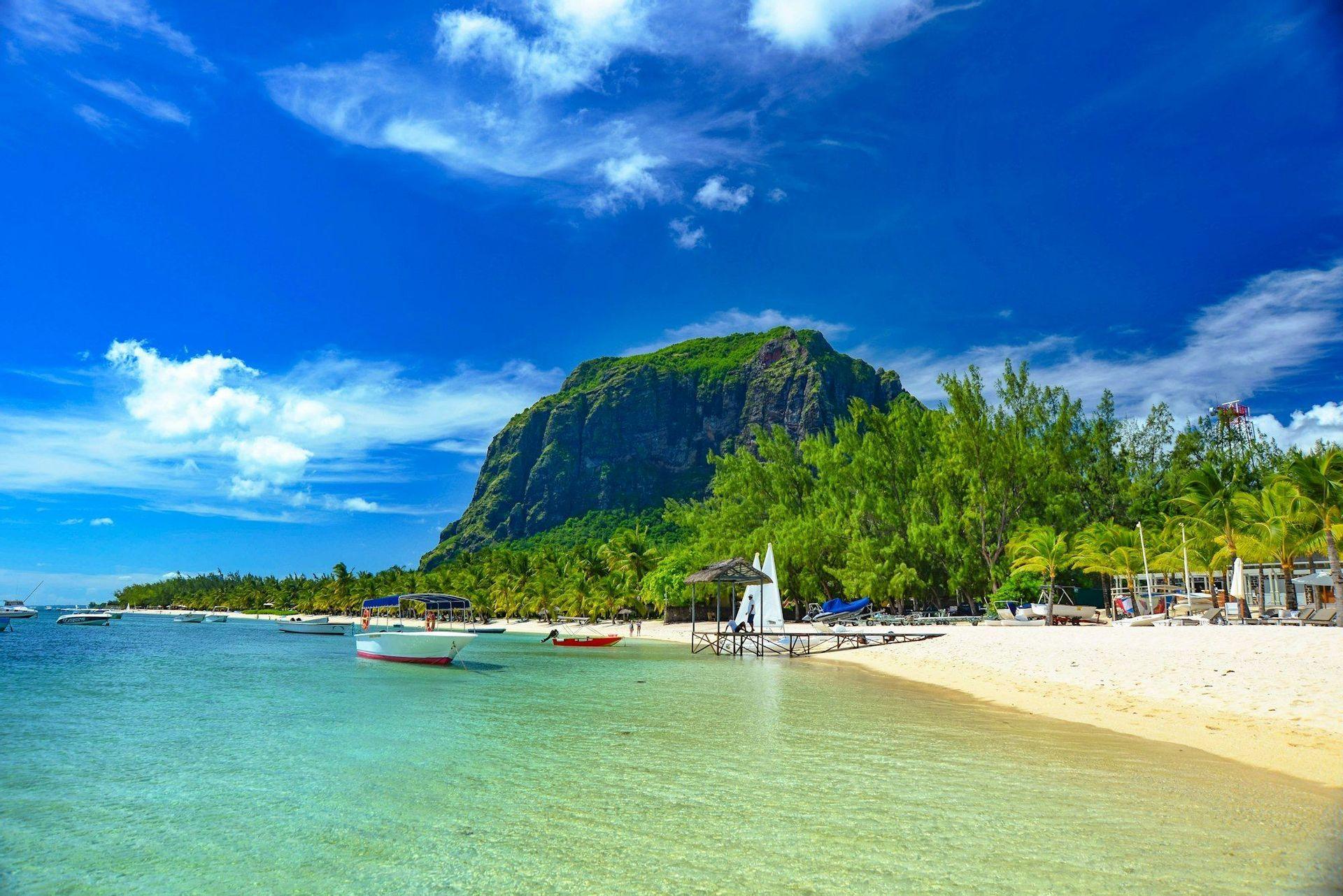 Una spiaggia tropicale con acque calme e turchesi e barche, sullo sfondo una grande montagna verde sotto un cielo azzurro brillante.