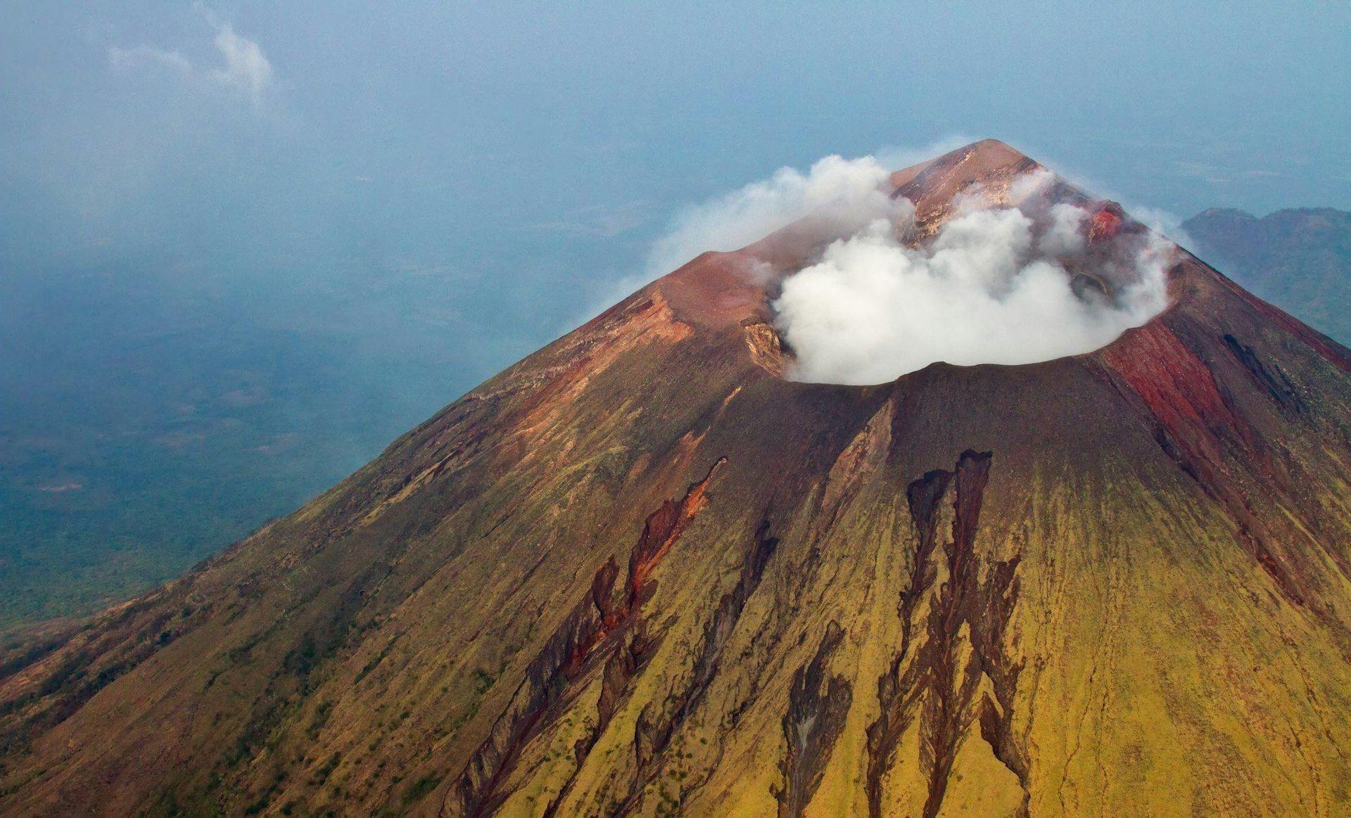 Vista aerea di un grande vulcano dai pendii colorati, con fumo bianco che si innalza dal suo cratere centrale.