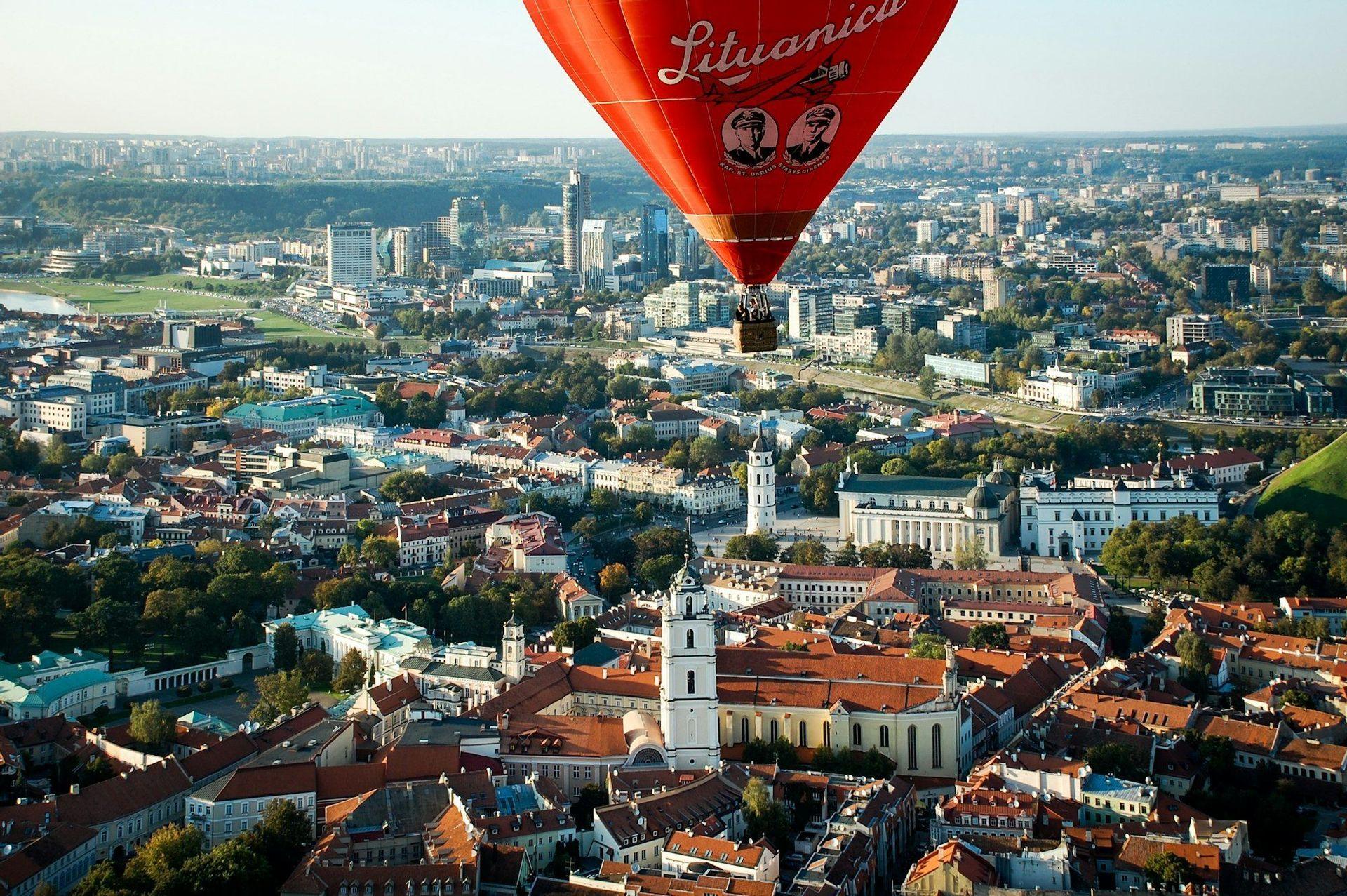 Ein großer roter Heißluftballon schwebt über einem historischen Stadtzentrum mit roten Ziegeldächern, im Hintergrund eine moderne Skyline.
