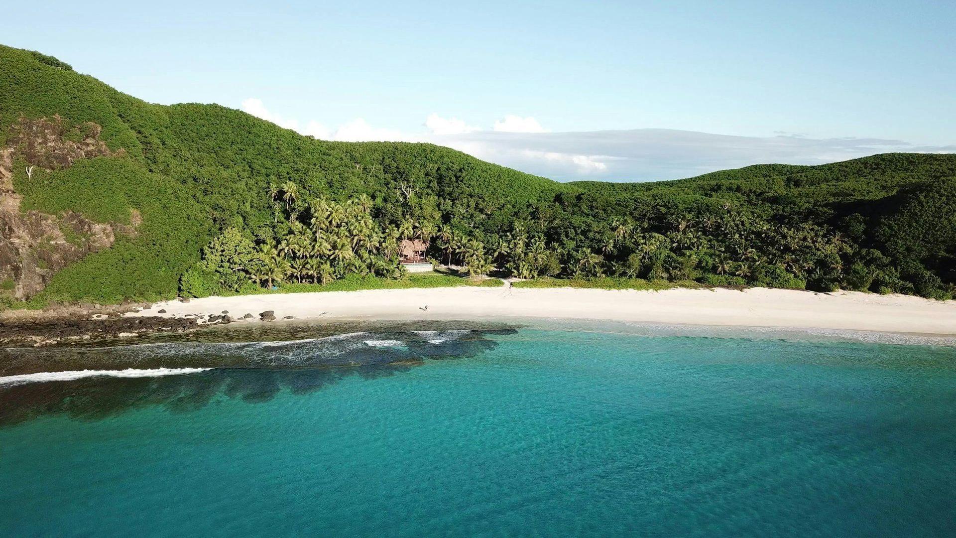 Una vista aérea de una playa solitaria de arena blanca con agua turquesa, respaldada por una exuberante colina verde cubierta de bosque tropical.