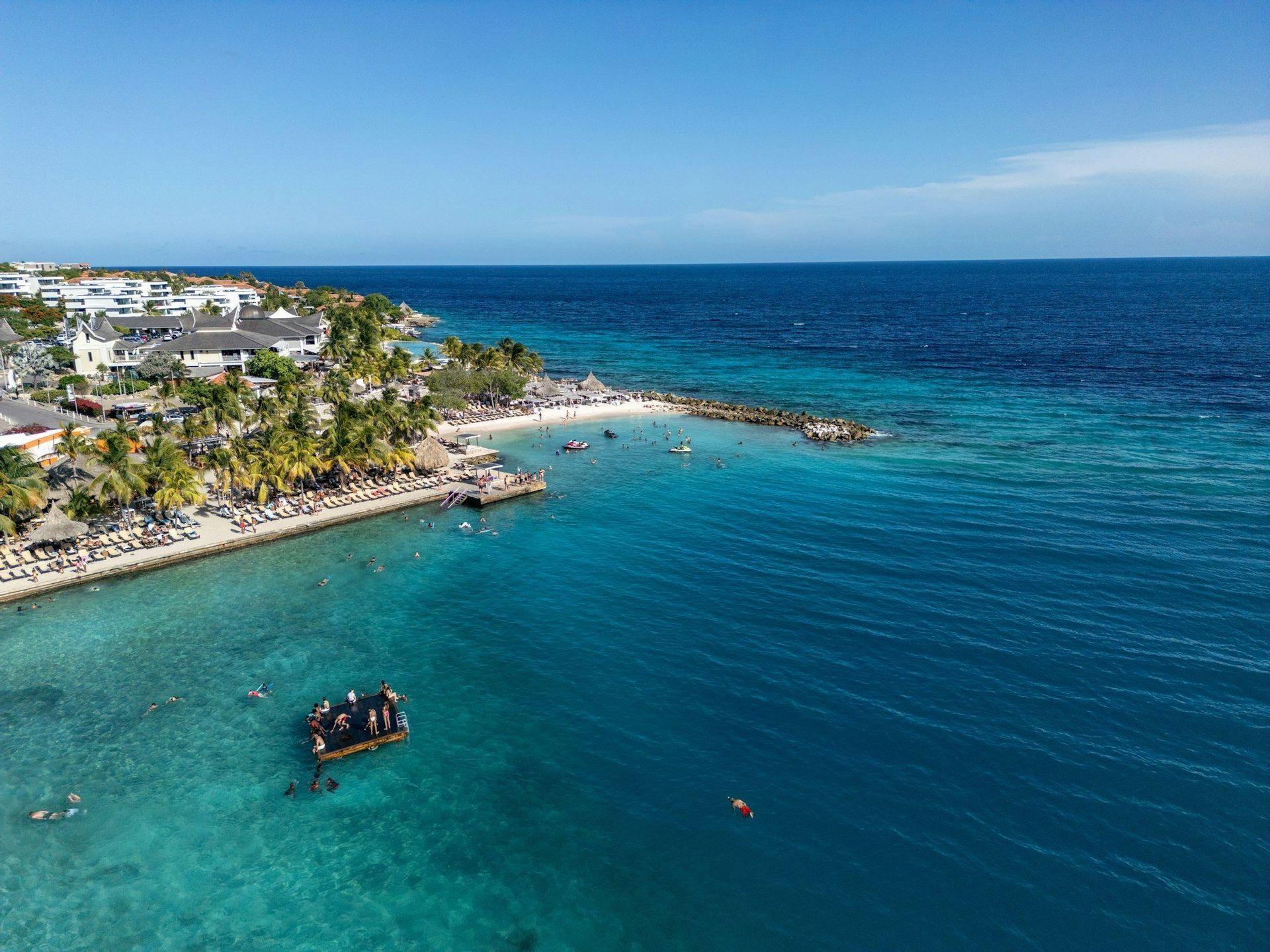 Vue aérienne d'un littoral tropical avec un complexe hôtelier en bord de mer, des palmiers et des personnes nageant dans la mer turquoise cristalline près d'une plateforme flottante.