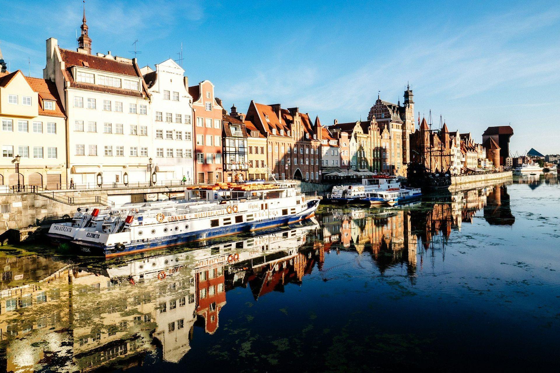 Une rangée de bâtiments historiques colorés et de bateaux amarrés le long d'un front de mer urbain, se reflétant dans l'eau calme par une journée ensoleillée.