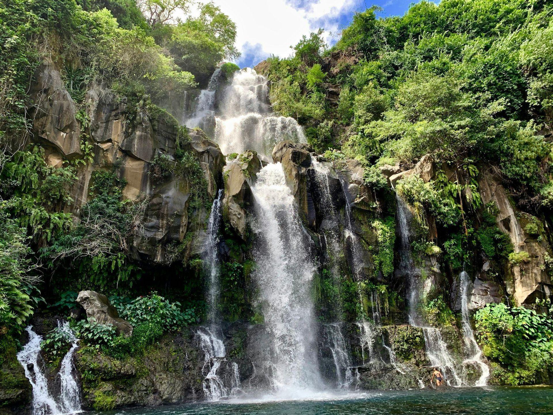 Une grande cascade dévale une falaise rocheuse couverte d'un feuillage vert luxuriant jusqu'à un bassin d'eau où une personne se baigne.