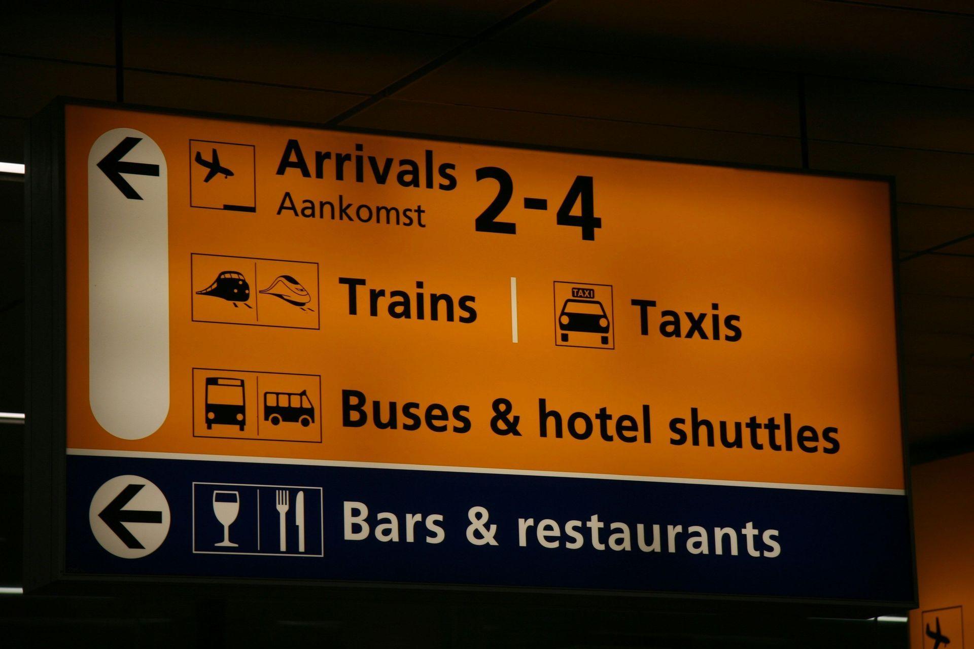 Un cartello aeroportuale illuminato indica gli arrivi, i treni, i taxi, gli autobus e i ristoranti, con pittogrammi.