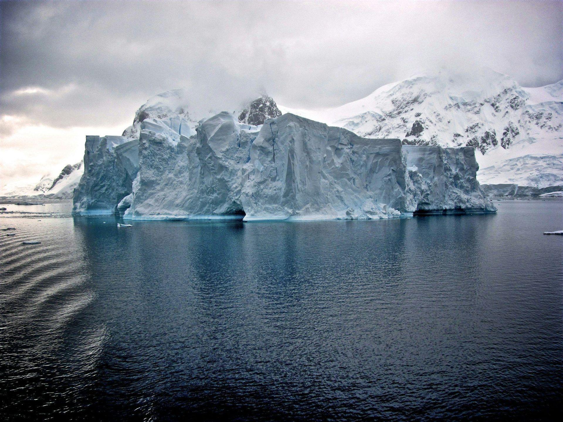 A large, craggy iceberg floats in deep blue water in front of snow-covered mountains under a cloudy sky.